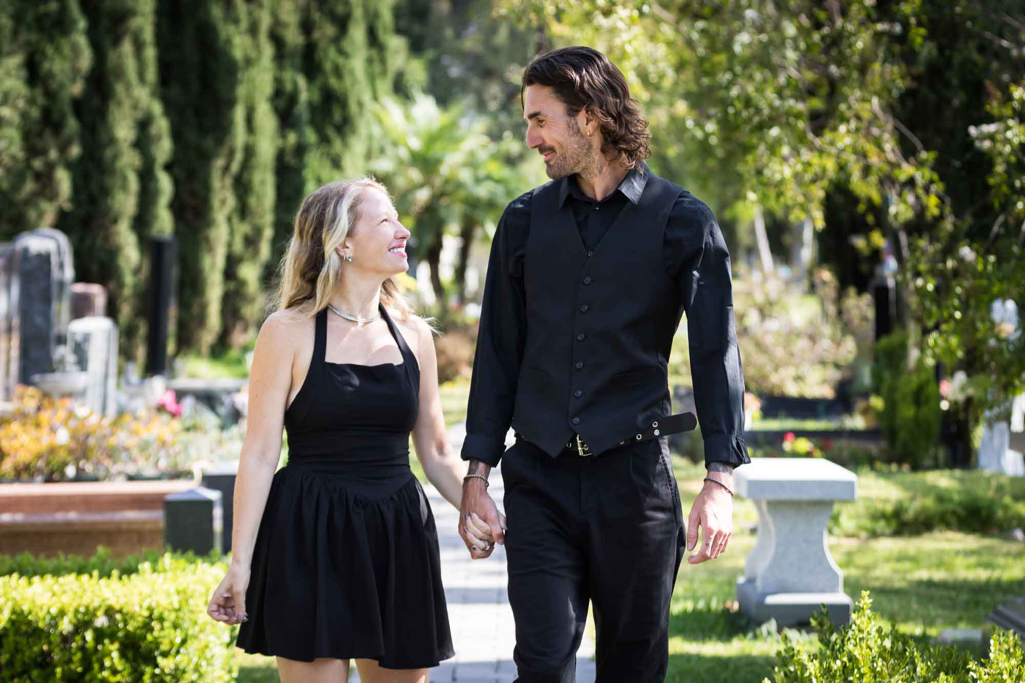 Man and woman wearing black outfits and holding hands while walking down Ancestral Forest walkway during a Hollywood Forever Cemetery engagement portrait session