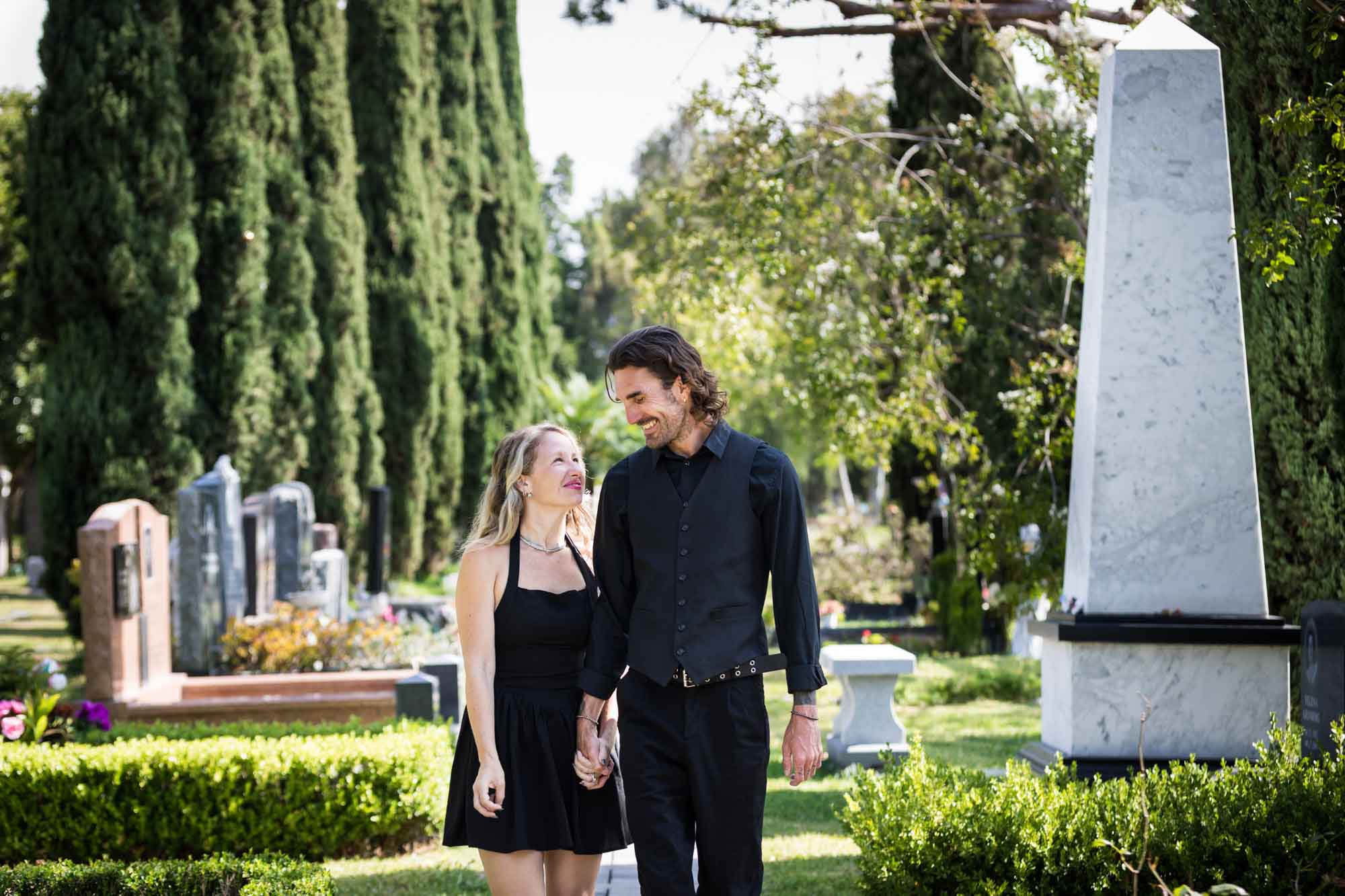 Man and woman wearing black outfits and holding hands while walking down Ancestral Forest walkway during a Hollywood Forever Cemetery engagement portrait session