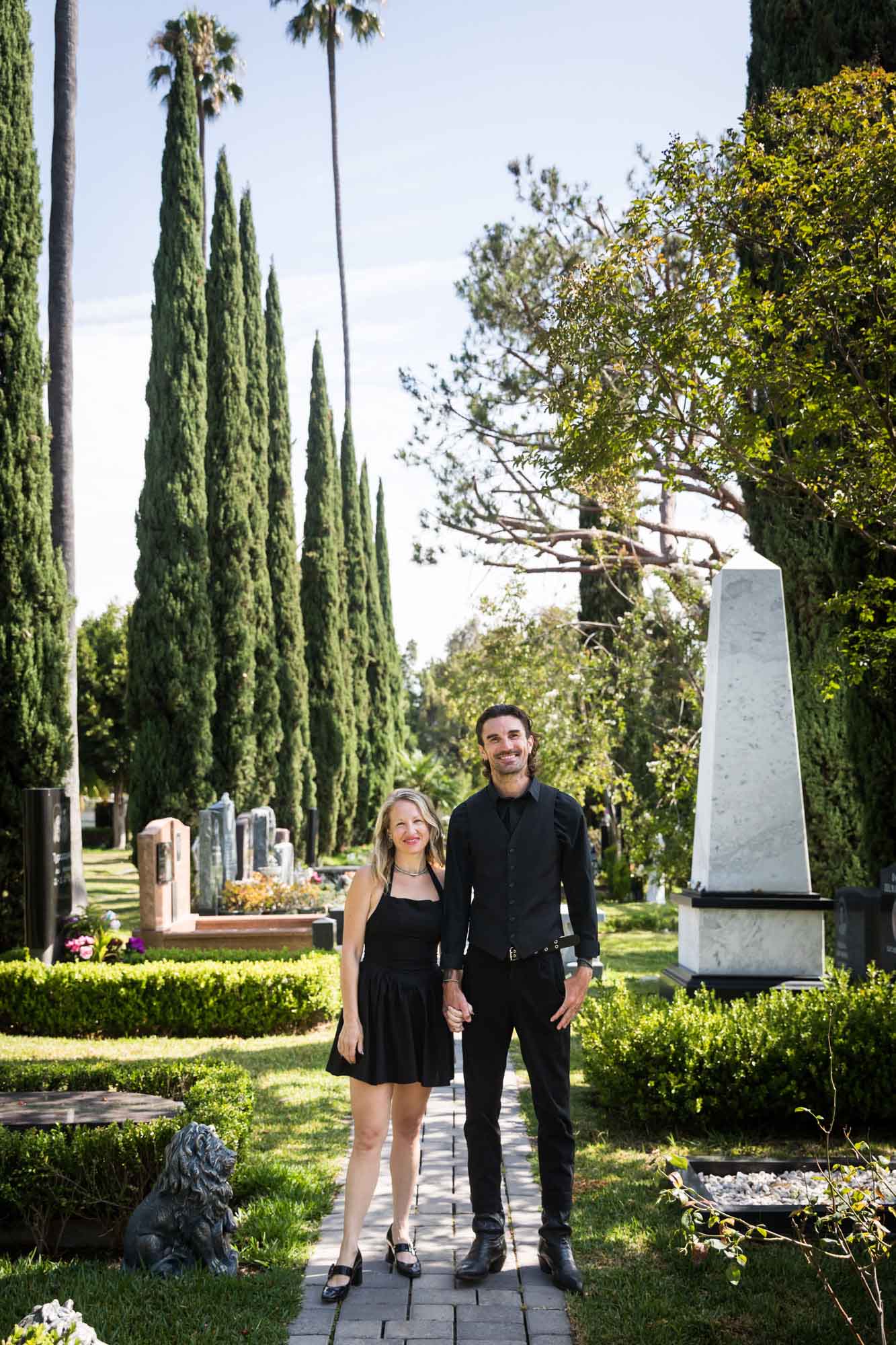 Man and woman wearing black outfits and holding hands while standing in Ancestral Forest walkway during a Hollywood Forever Cemetery engagement portrait session