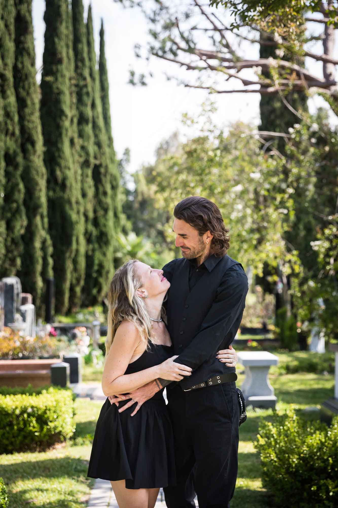 Man and woman looking at each other and hugging in Ancestral Forest walkway during a Hollywood Forever Cemetery engagement portrait session