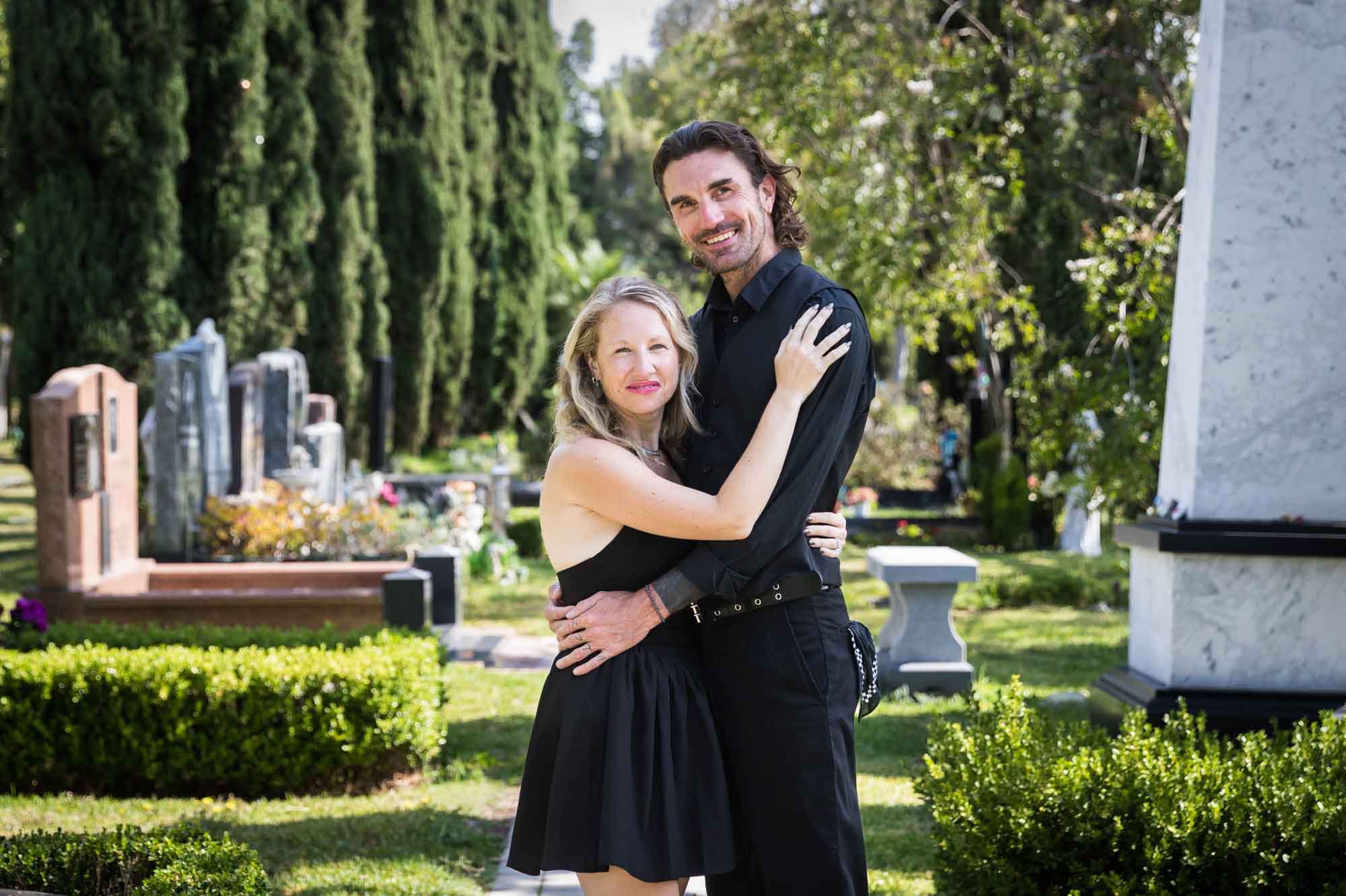 Man and woman hugging in front of tombstones and trees during a Hollywood Forever Cemetery engagement portrait session