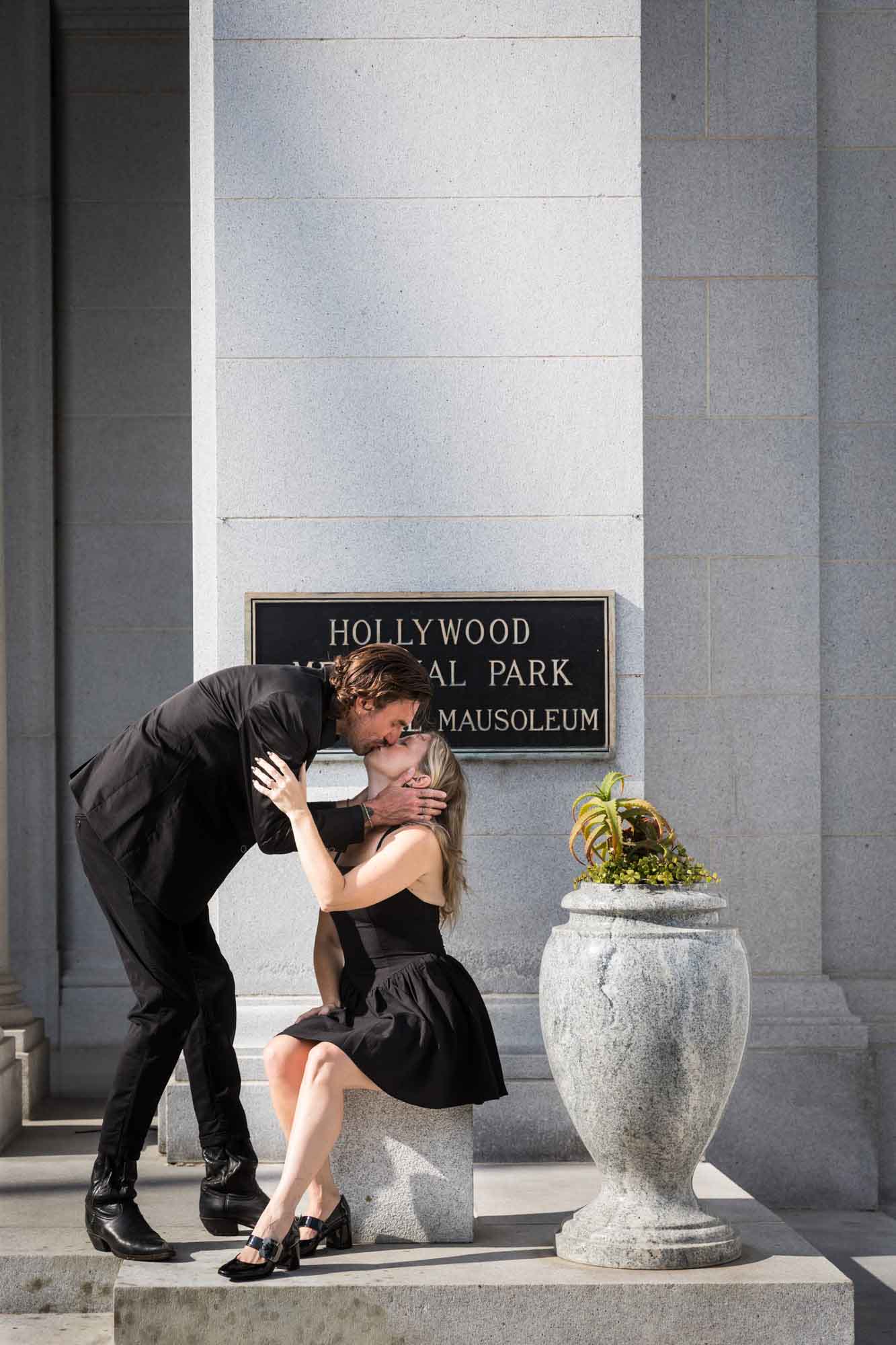 Man reaching down to kiss woman sitting on stone pillar next to urn outside Cathedral Mausoleum during a Hollywood Forever Cemetery engagement portrait session