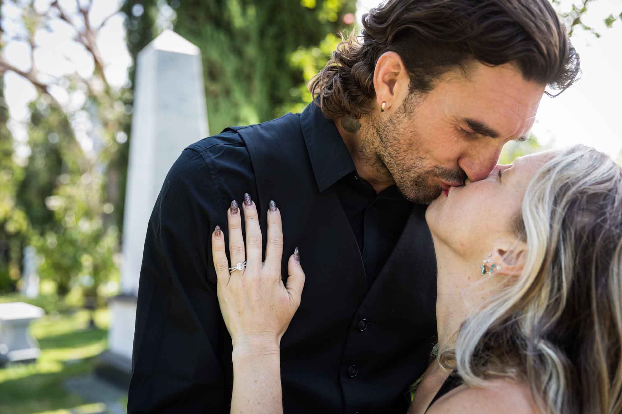 Close up of man and woman kissing with woman's hand showing engagement ring on man's shoulder during a Hollywood Forever Cemetery engagement portrait session