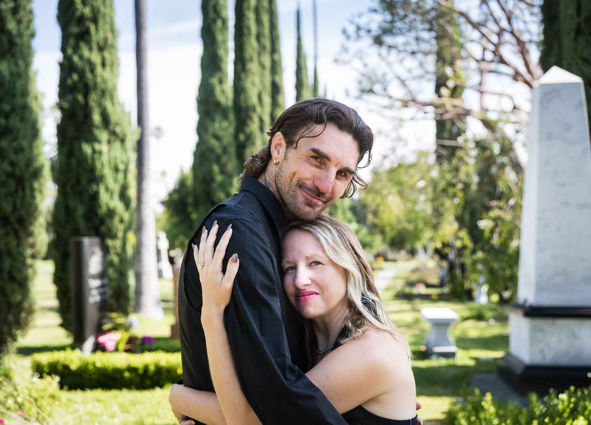 Man and woman hugging in front of tombstones and trees during a Hollywood Forever Cemetery engagement portrait session