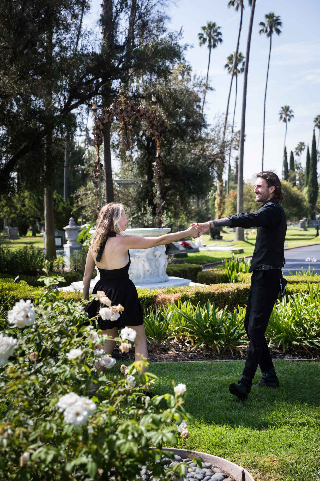Man and woman dancing behind rose bush and in front of antique well during a Hollywood Forever Cemetery engagement portrait session