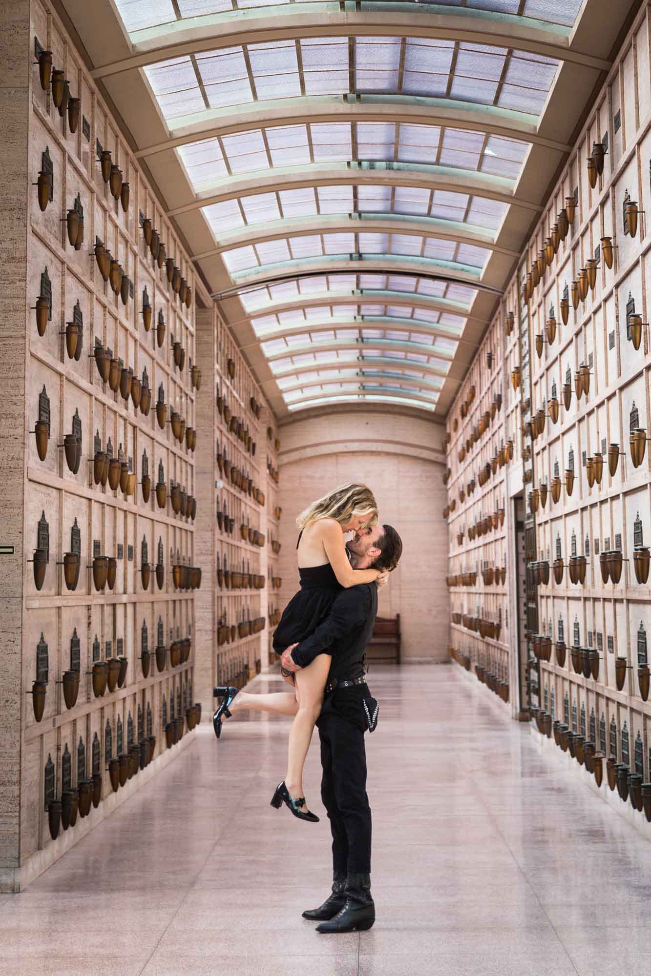 Man lifting woman up in the air in crypt hallway in the Abbey of the Psalms during a Hollywood Forever Cemetery engagement portrait session
