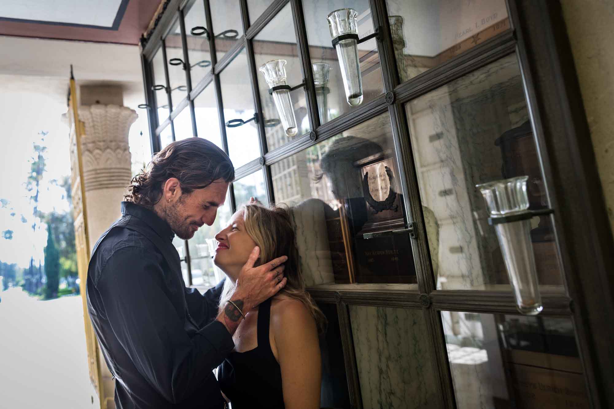 Man holding woman's head while resting against glass wall during a Hollywood Forever Cemetery engagement portrait session