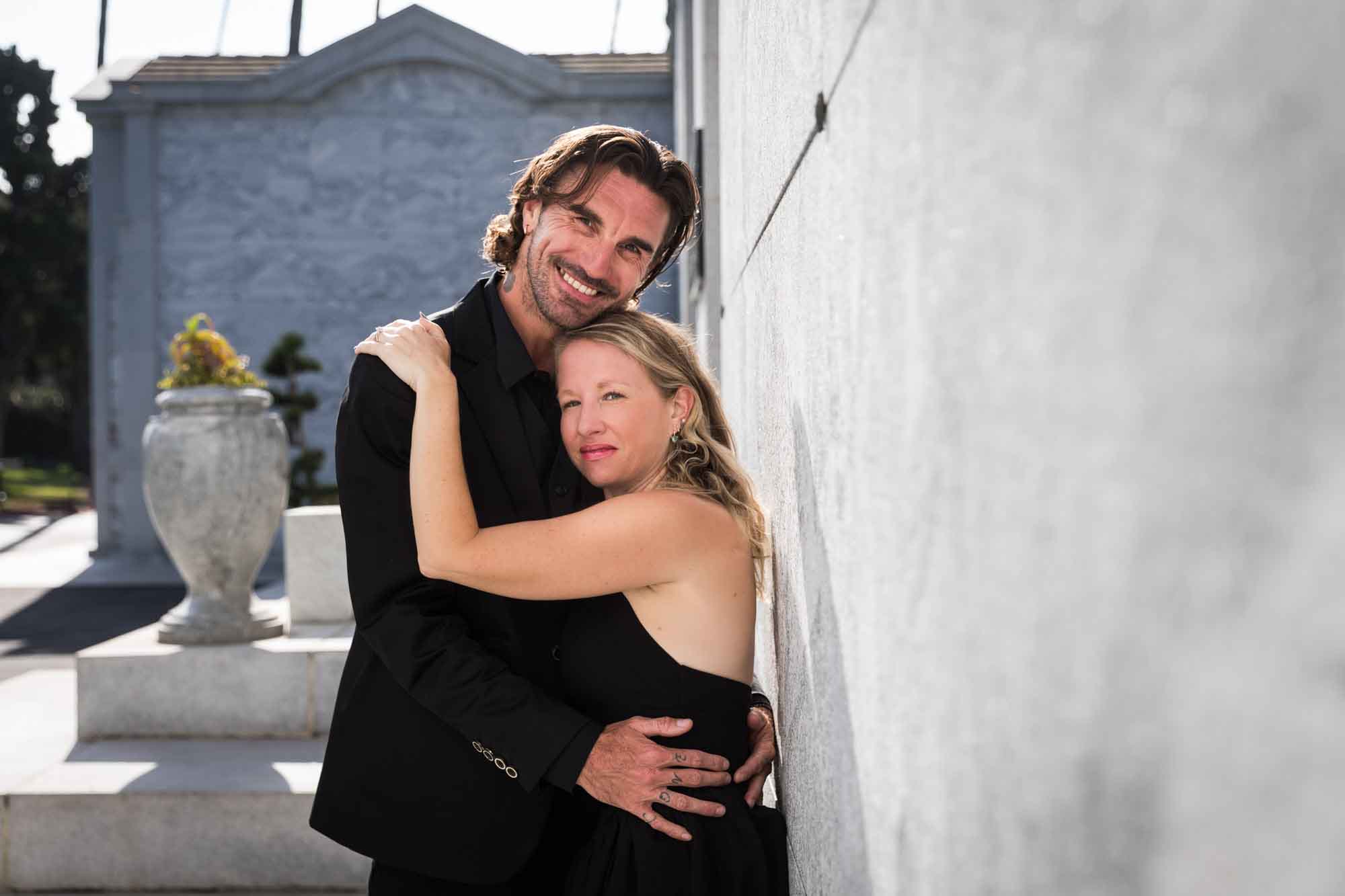 Man and woman wearing black clothes hugging next to wall during a Hollywood Forever Cemetery engagement portrait session