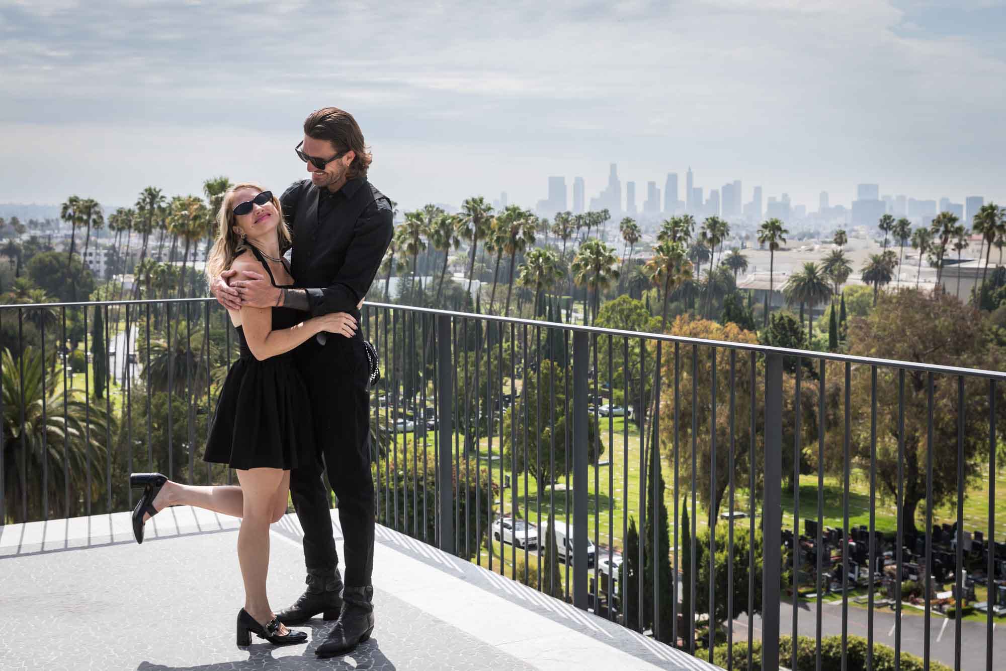 Man hugging woman holding leg up in air on rooftop with downtown Los Angeles skyline in background during a Hollywood Forever Cemetery engagement portrait session