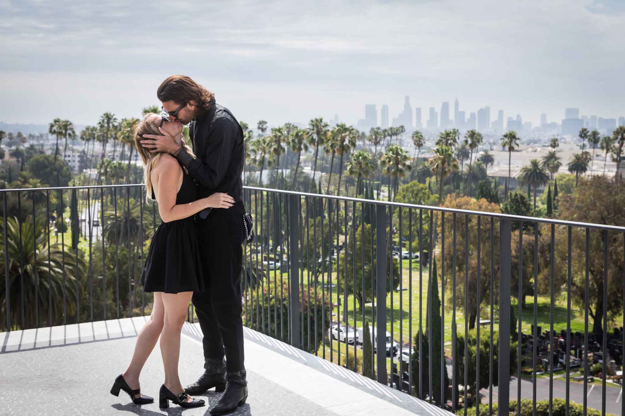 Man holding woman's facing while kissing her on rooftop with black railing and Los Angeles skyline in background during a Hollywood Forever Cemetery engagement portrait session
