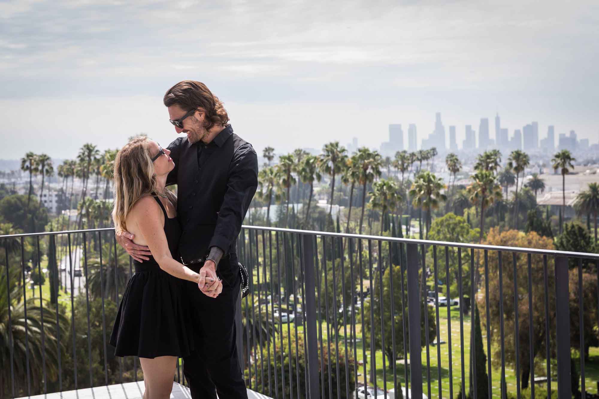 Man and woman wearing black outfits dancing close on rooftop with black railing and downtown Los Angeles skyline in background during a Hollywood Forever Cemetery engagement portrait session