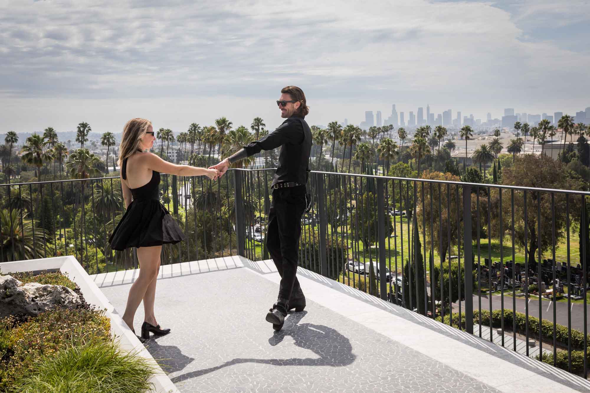 Man and woman wearing black outfits dancing on rooftop with black railing and downtown Los Angeles skyline and clouds in background during a Hollywood Forever Cemetery engagement portrait session