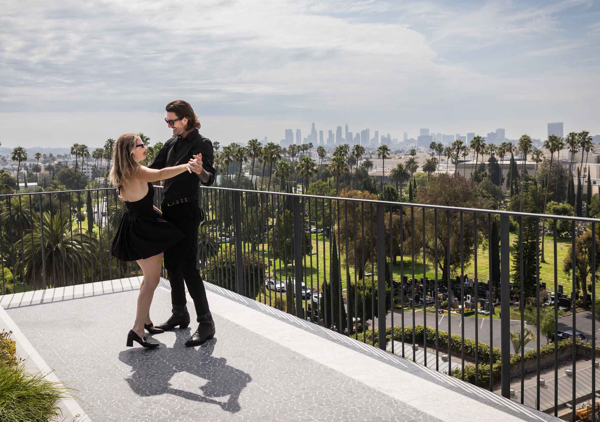 Man and woman dancing on rooftop with black railing and Los Angeles downtown background in background during a Hollywood Forever Cemetery engagement portrait session