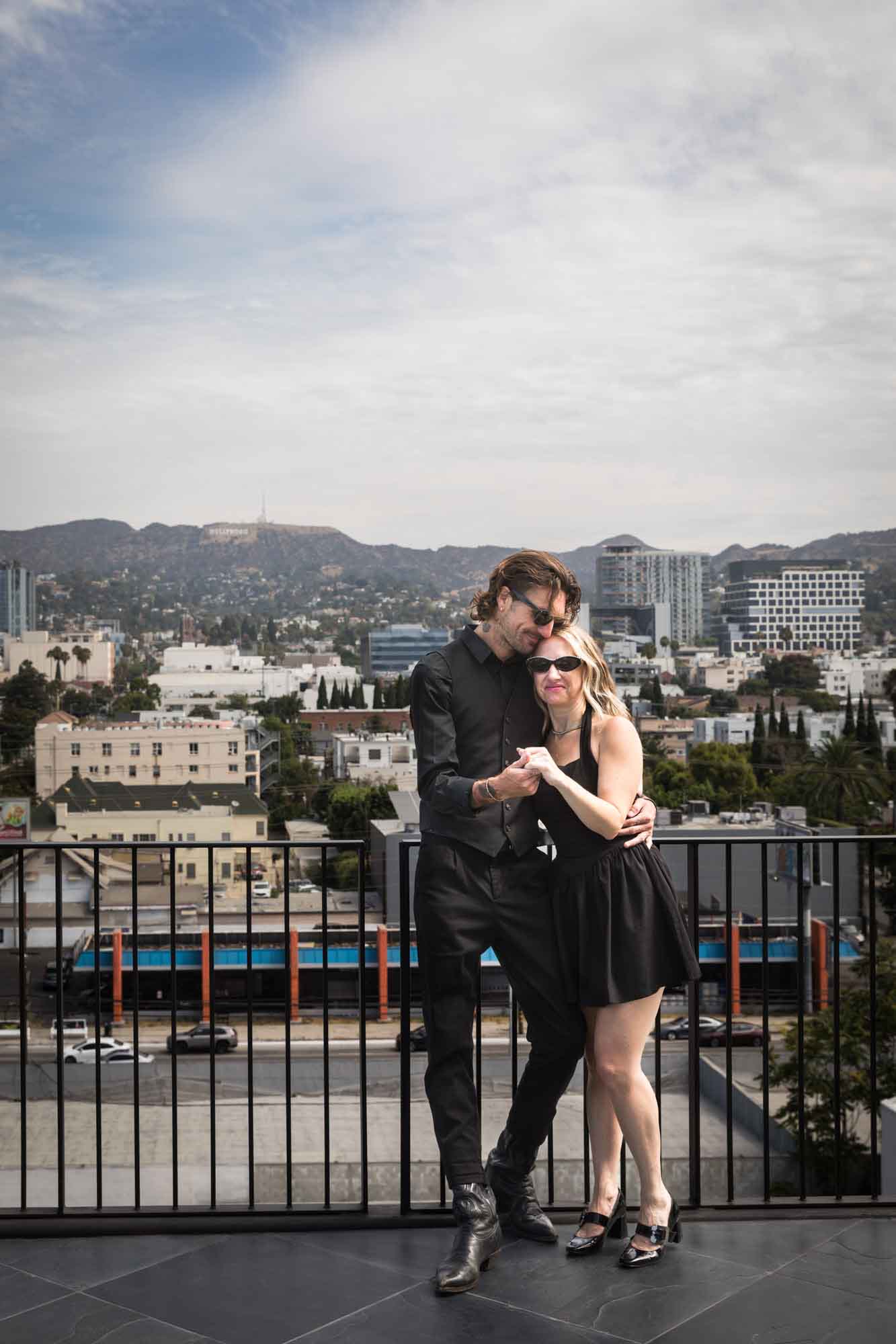 Man and woman holding hands in front of railing on rooftop with Los Angeles skyline in background during a Hollywood Forever Cemetery engagement portrait session