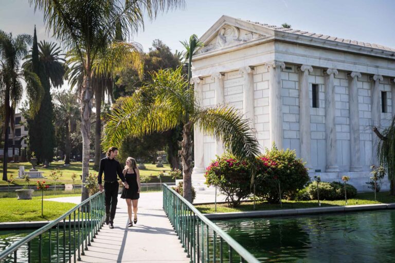Hollywood Forever Cemetery engagement portrait of man and woman walking down pathway with Clark Mausoleum and lake in background