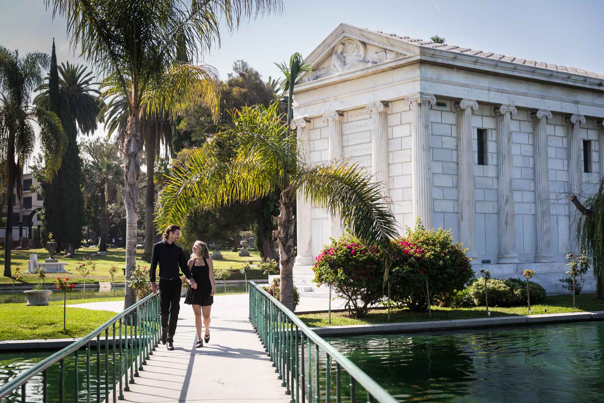 Hollywood Forever Cemetery engagement portrait of man and woman walking down pathway with Clark Mausoleum and lake in background