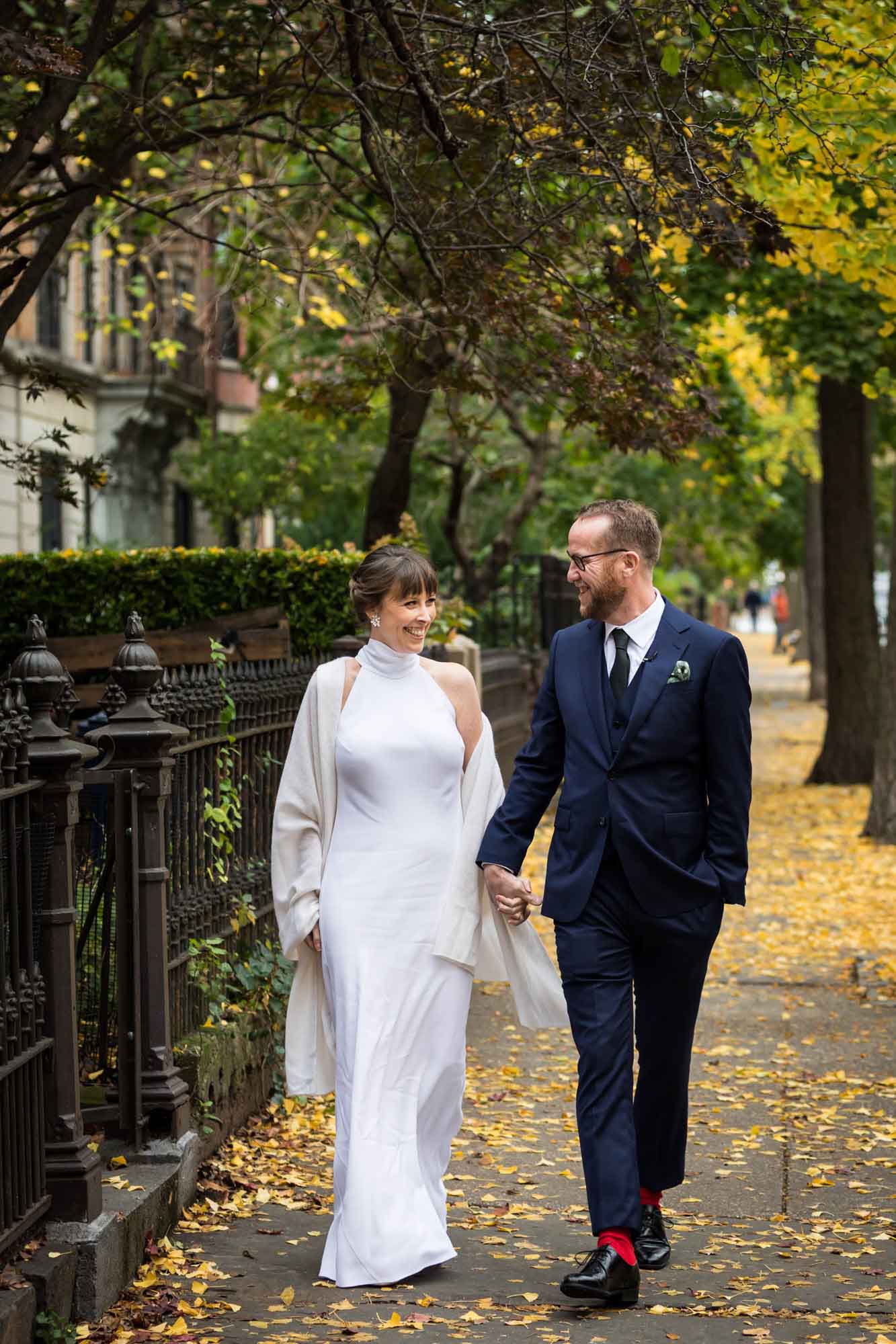 Bride and groom holding hands and walking down tree-lined sidewalk with brownstones in the background for an article on how to get a marriage license in Los Angeles
