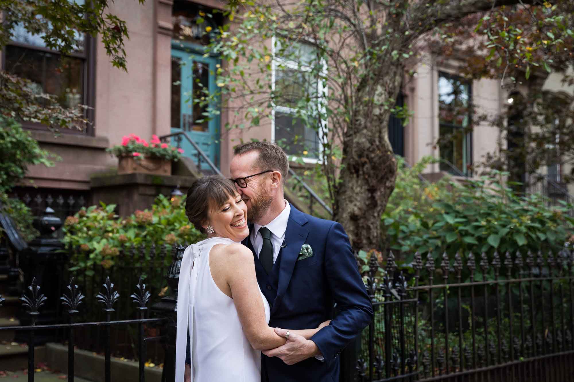 Groom kissing bride on the cheek on city sidewalk with trees and brownstones in the background for an article on how to get a marriage license in Los Angeles