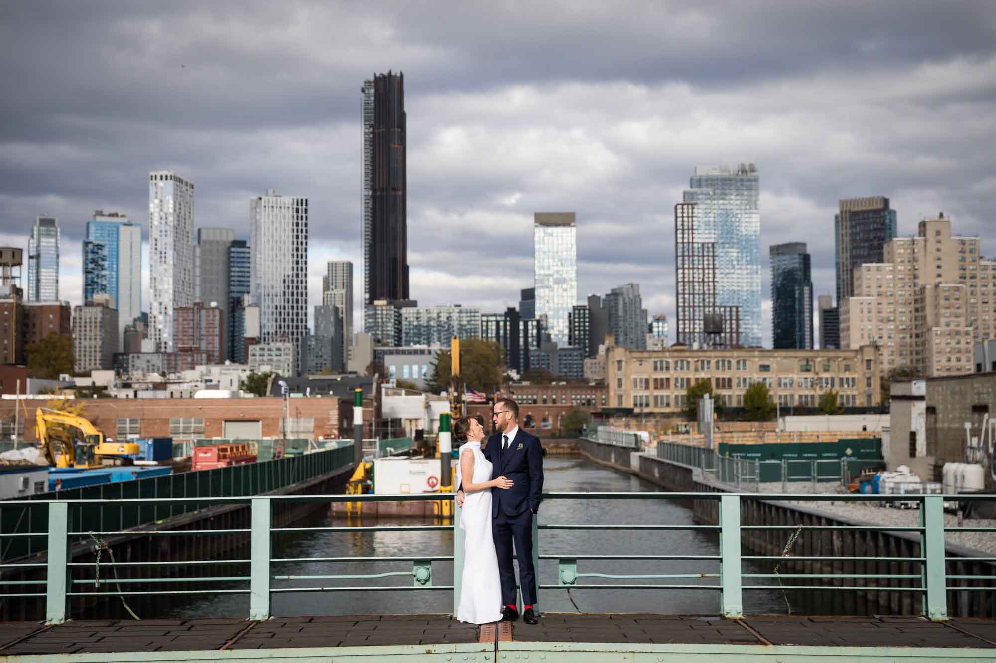 Bride and groom standing close together on bridge across water with city skyline in the background for an article on how to get a marriage license in Los Angeles