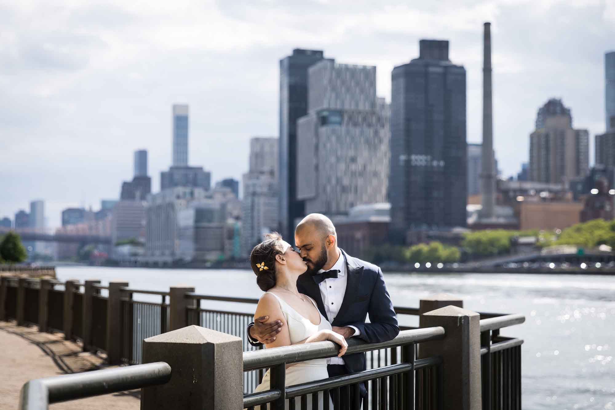 Bride and groom kissing while standing against railing with waterfront and city skyline in background for an article on how to get a marriage license in Los Angeles