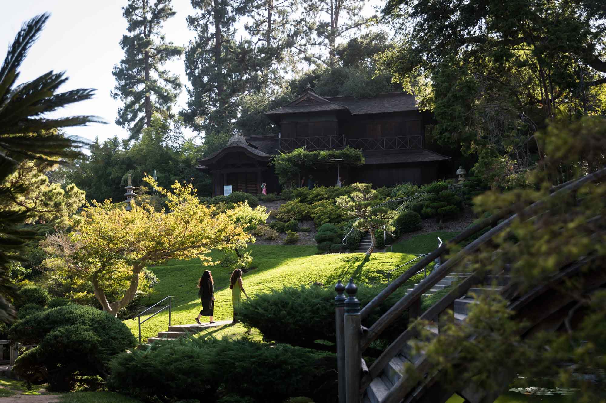 Two women walking along pathway with wooden bridge and wooden building in background in Japanese Garden for a Huntington Gardens photo shoot guide article
