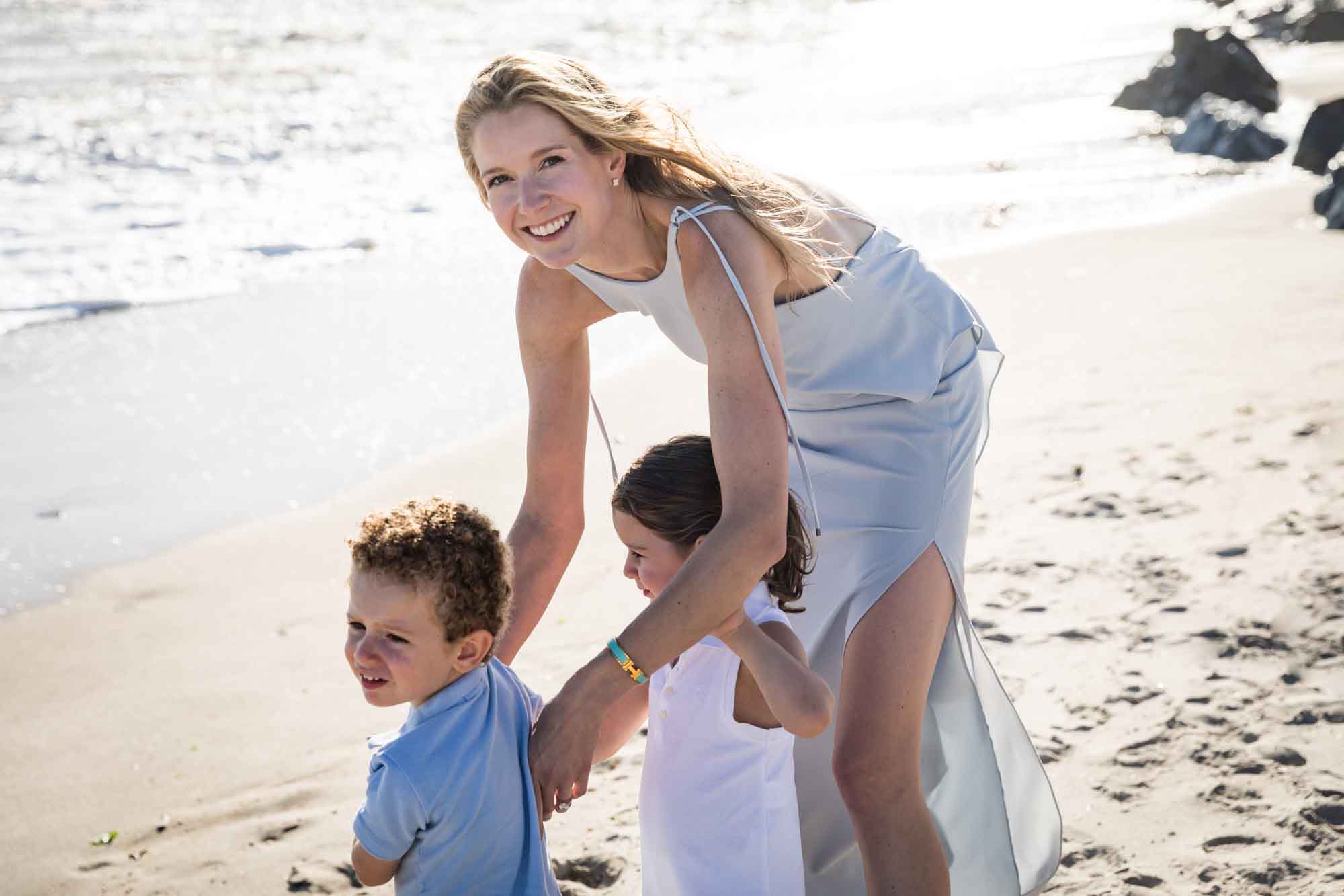 Mother bending down in sand to little boy and girl standing in front of beach and ocean for an article on La Jolla Cove family photos