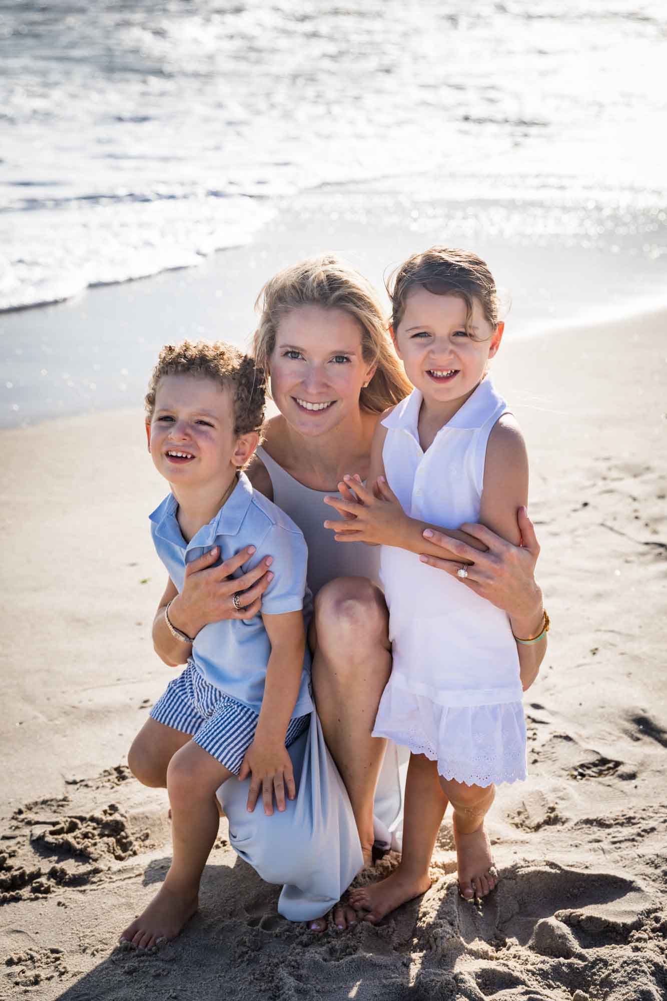 Mother crouched down in sand holding little boy and girl standing in front of beach and ocean for an article on La Jolla Cove family photos