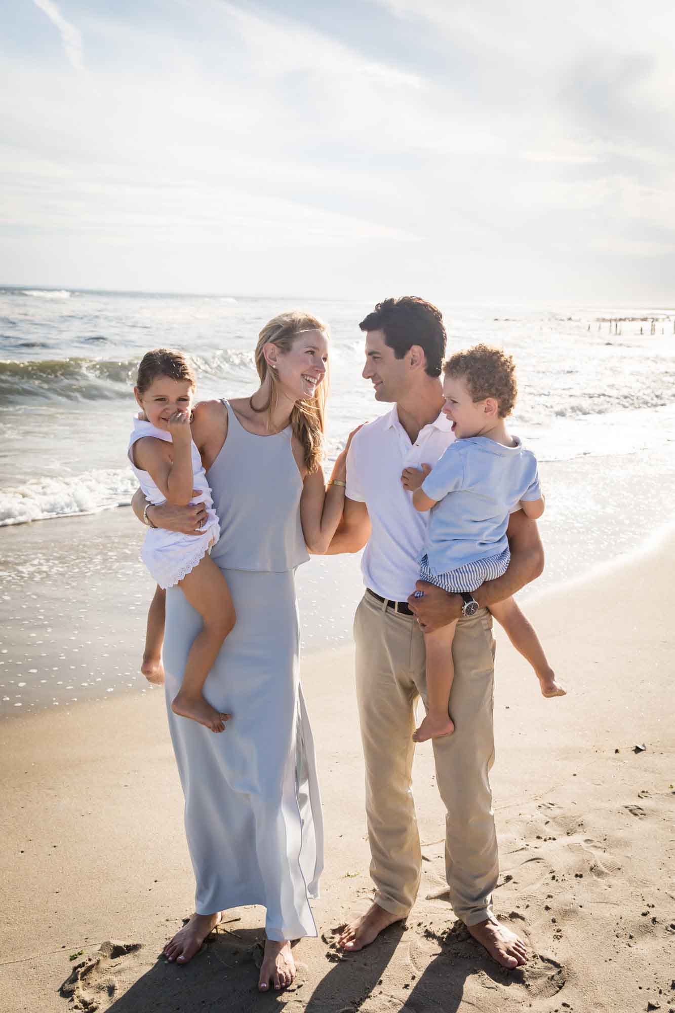 Mother and father standing barefoot on sand holding little boy and girl with ocean and waves in the background for an article on La Jolla Cove family photos