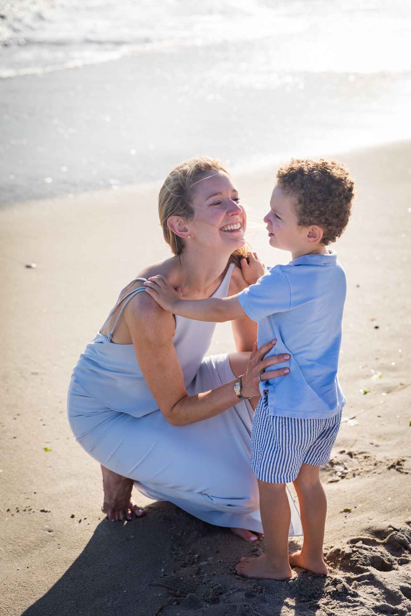 Mother crouched down looking at little boy wearing blue shirt with hands on mother's shoulders standing in sand at the beach for an article on La Jolla Cove family photos