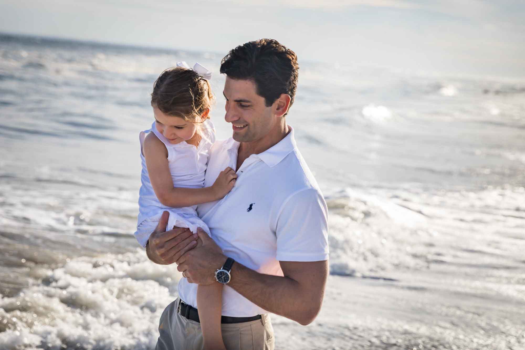 Father with dark hair holding little girl in his arms with ocean and waves in the background for an article on La Jolla Cove family photos