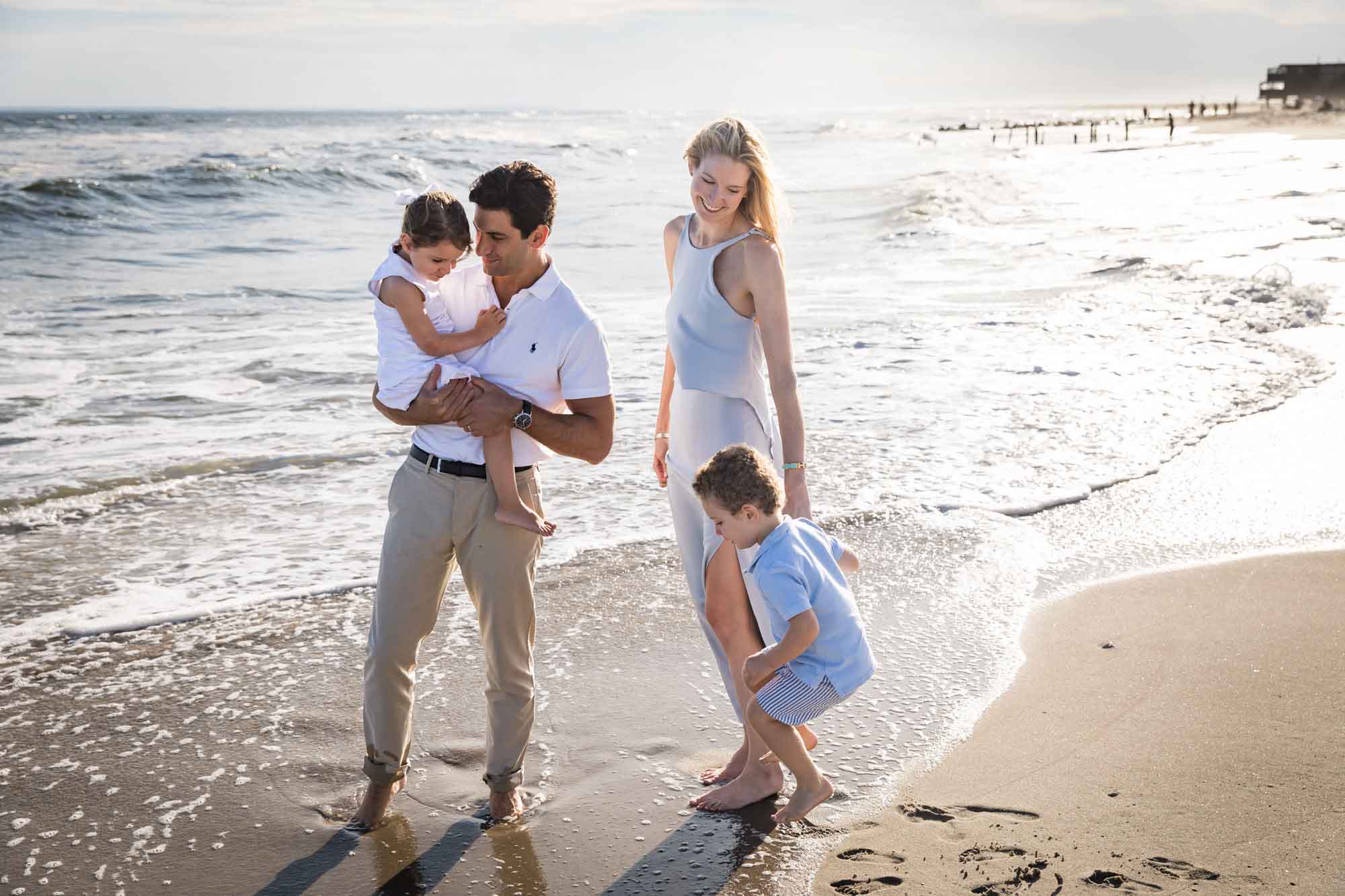Father holding little girl and mother holding hands with little boy while standing in the surf at the beach for an article on La Jolla Cove family photos