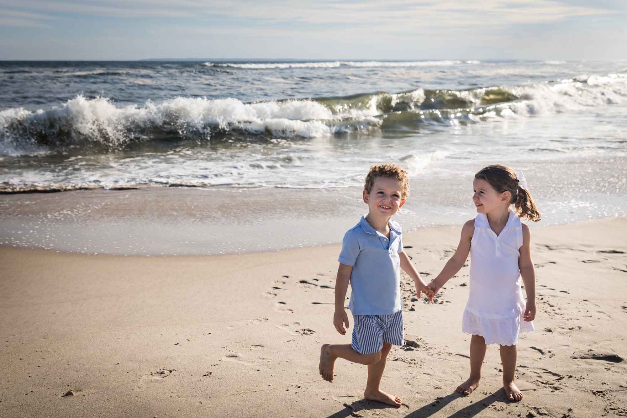 Little boy and girl holding hands while standing on the beach in front of the ocean with waves crashing for an article on La Jolla Cove family photos