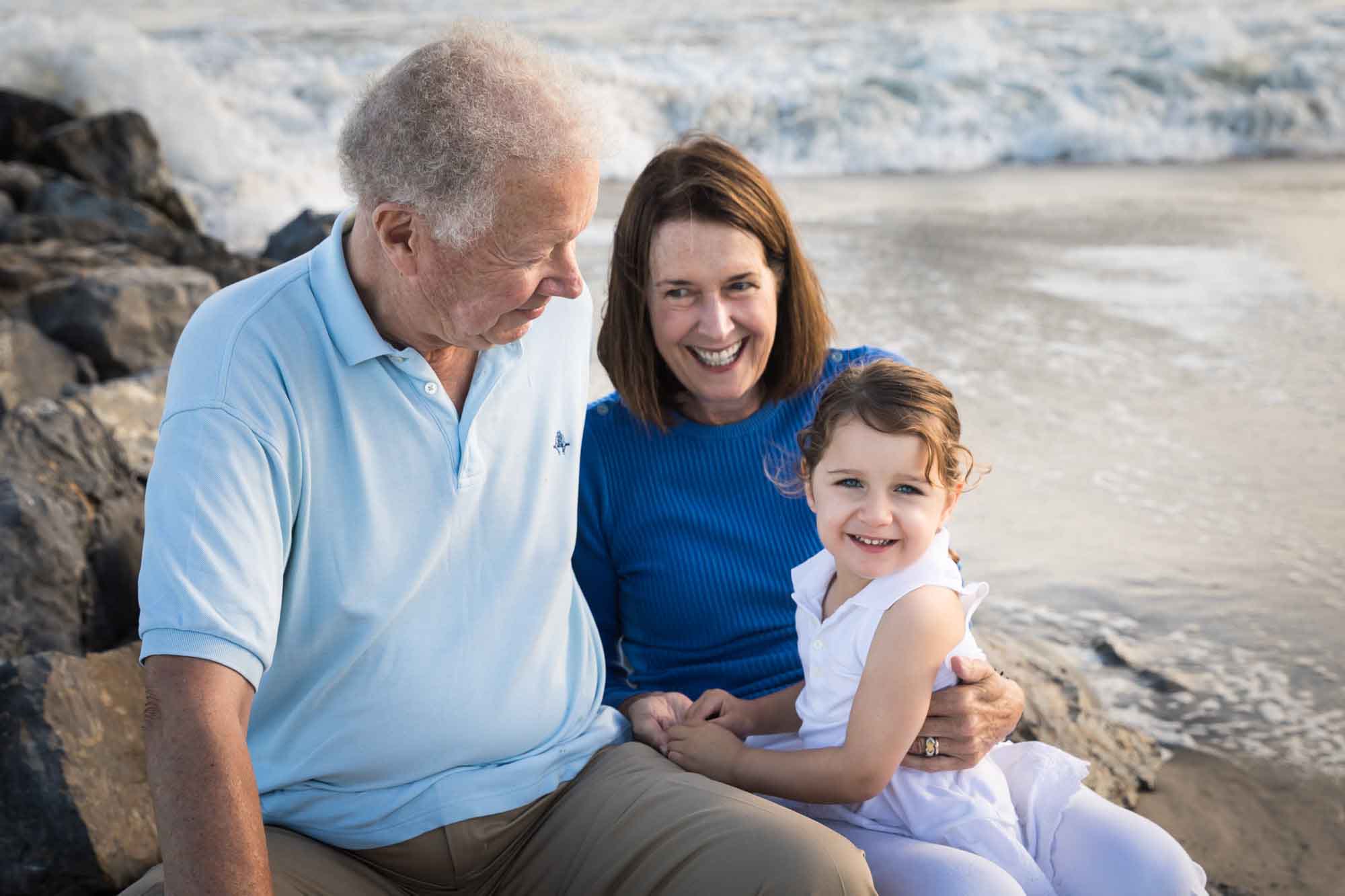 Grandparents holding little girl while sitting on rocks in front of the ocean for an article on La Jolla Cove family photos