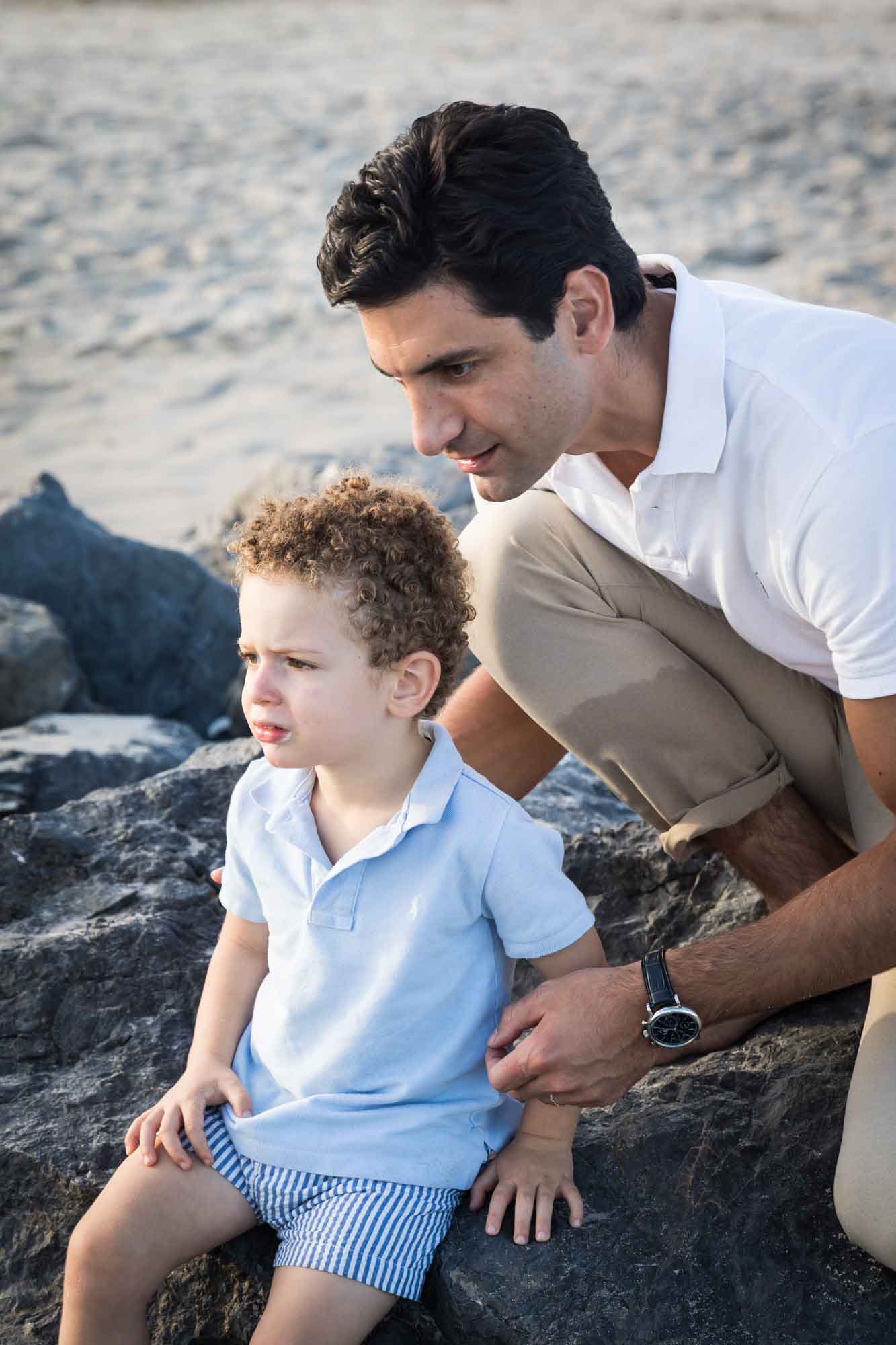 Father with dark brown hair looking down with little boy wearing blue and white striped shorts while sitting on rocks for an article on La Jolla Cove family photos