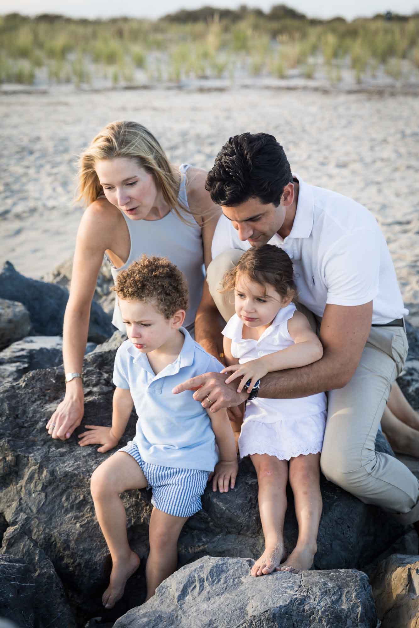 Mother and father holding little boy and girl while sitting on rocks pointing to something on the ground for an article on La Jolla Cove family photos