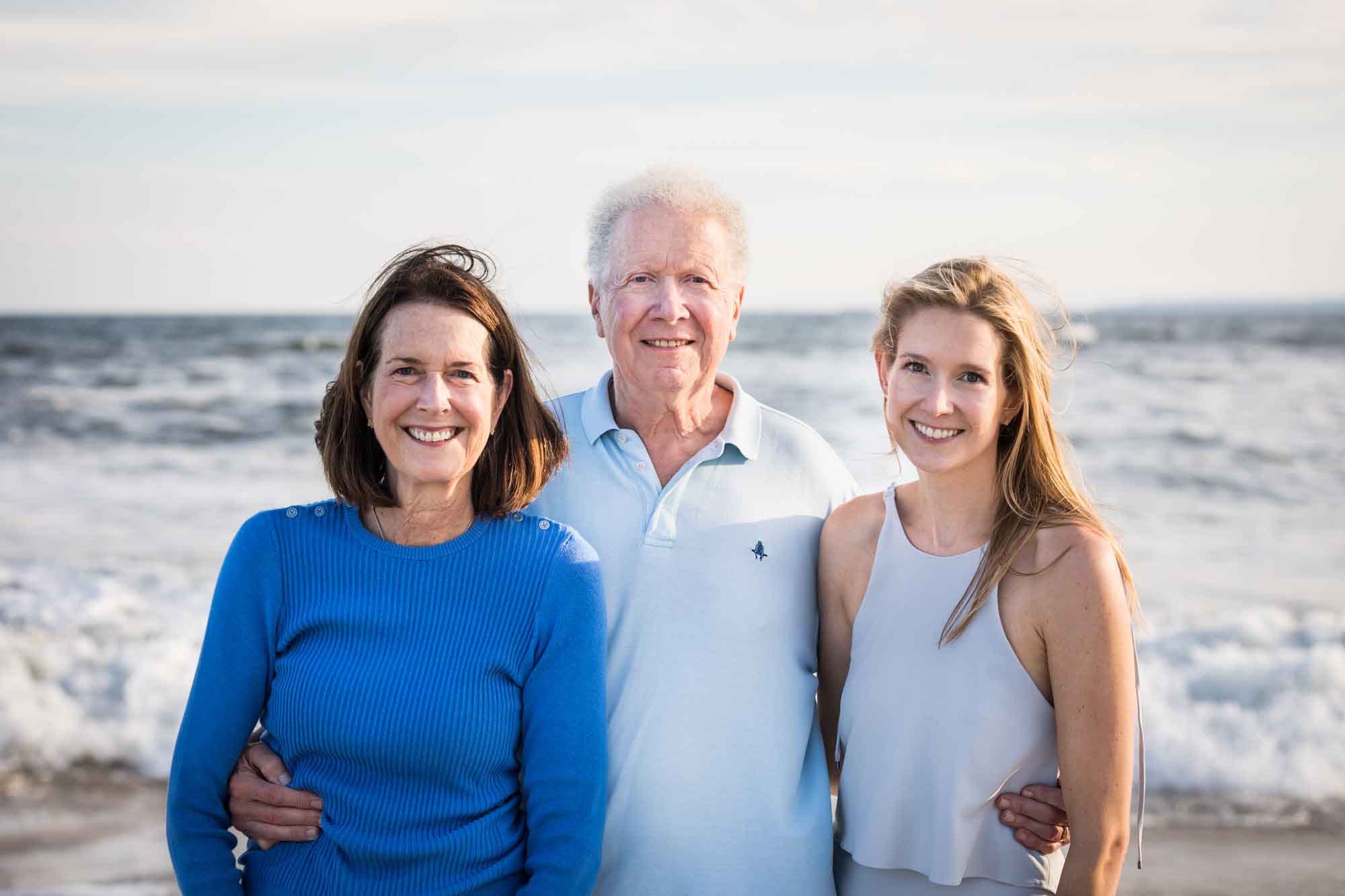 Man standing in between woman wearing blue shirt and younger woman wearing sleeveless blouse in front of ocean for an article on La Jolla Cove family photos
