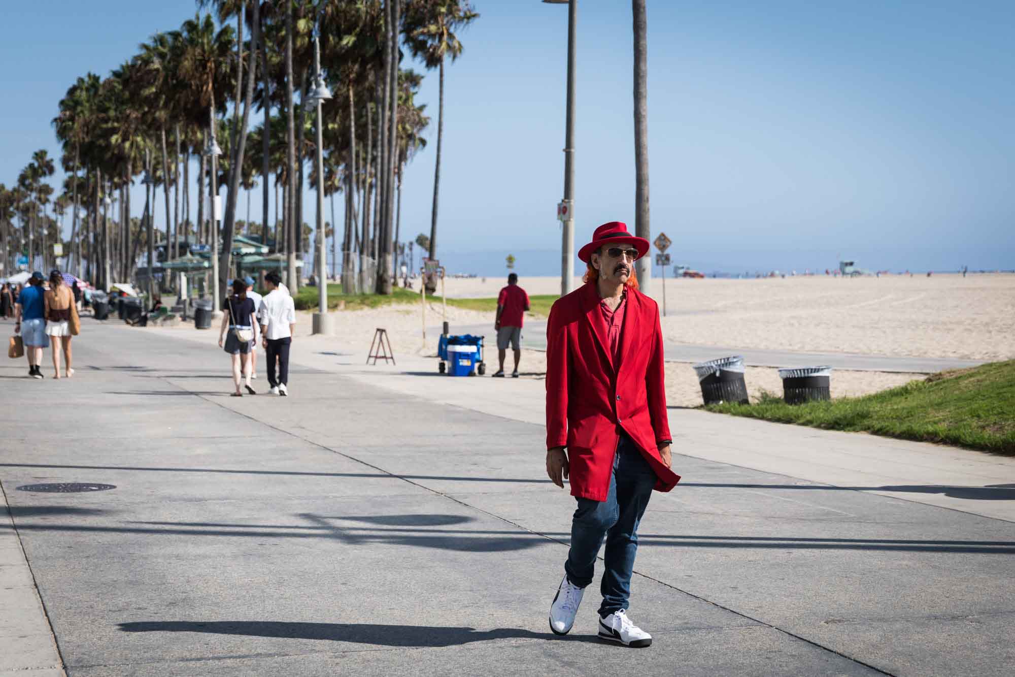 An eccentric man wearing a long red coat, red hat, and sunglasses walking along the boardwalk in Venice Beach for an article on Venice Canals engagement photos