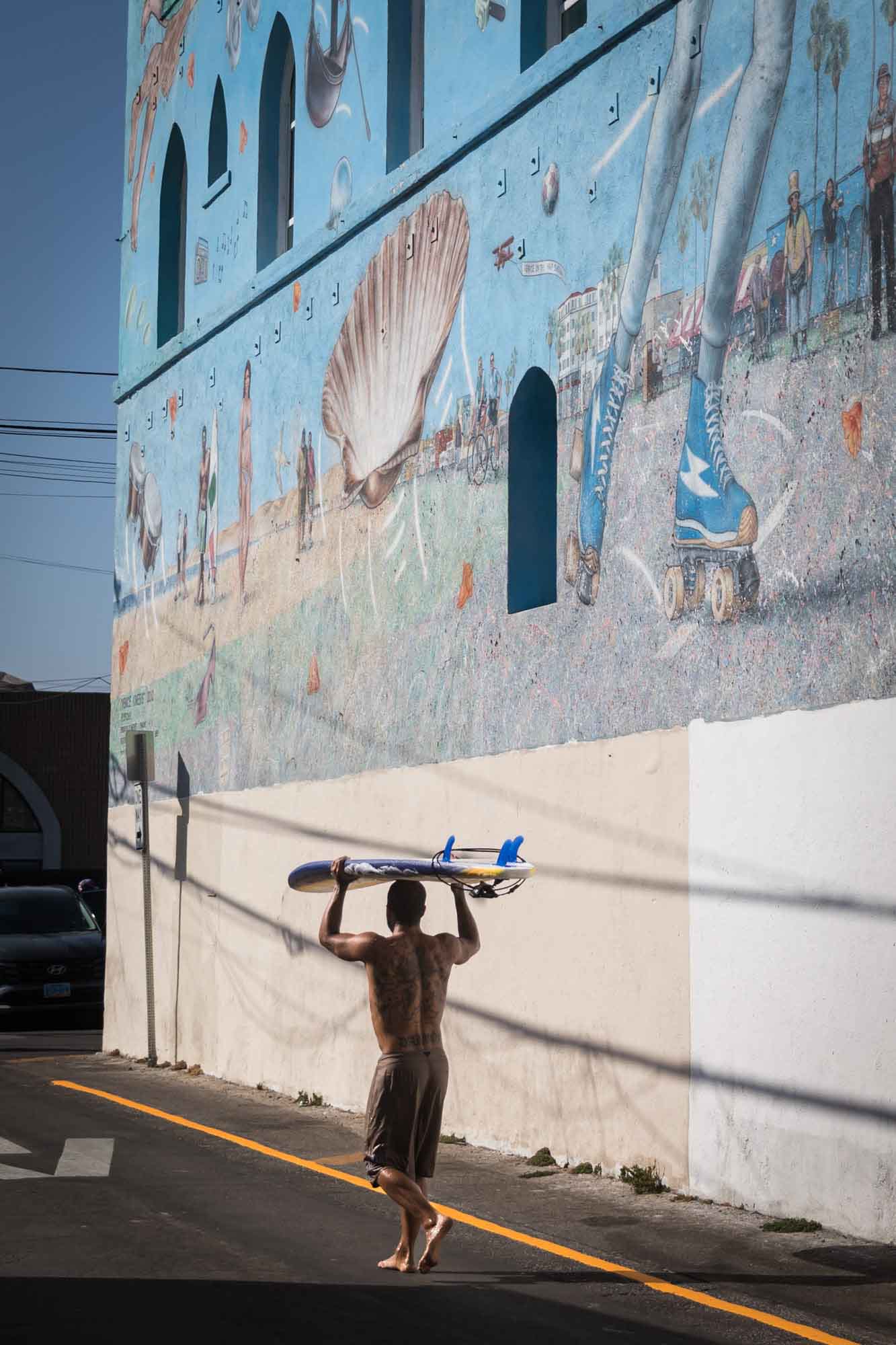 Male surfer walking barefoot and holding surfboard above his head in front of wall with colorful mural for an article on Venice Canals engagement photos