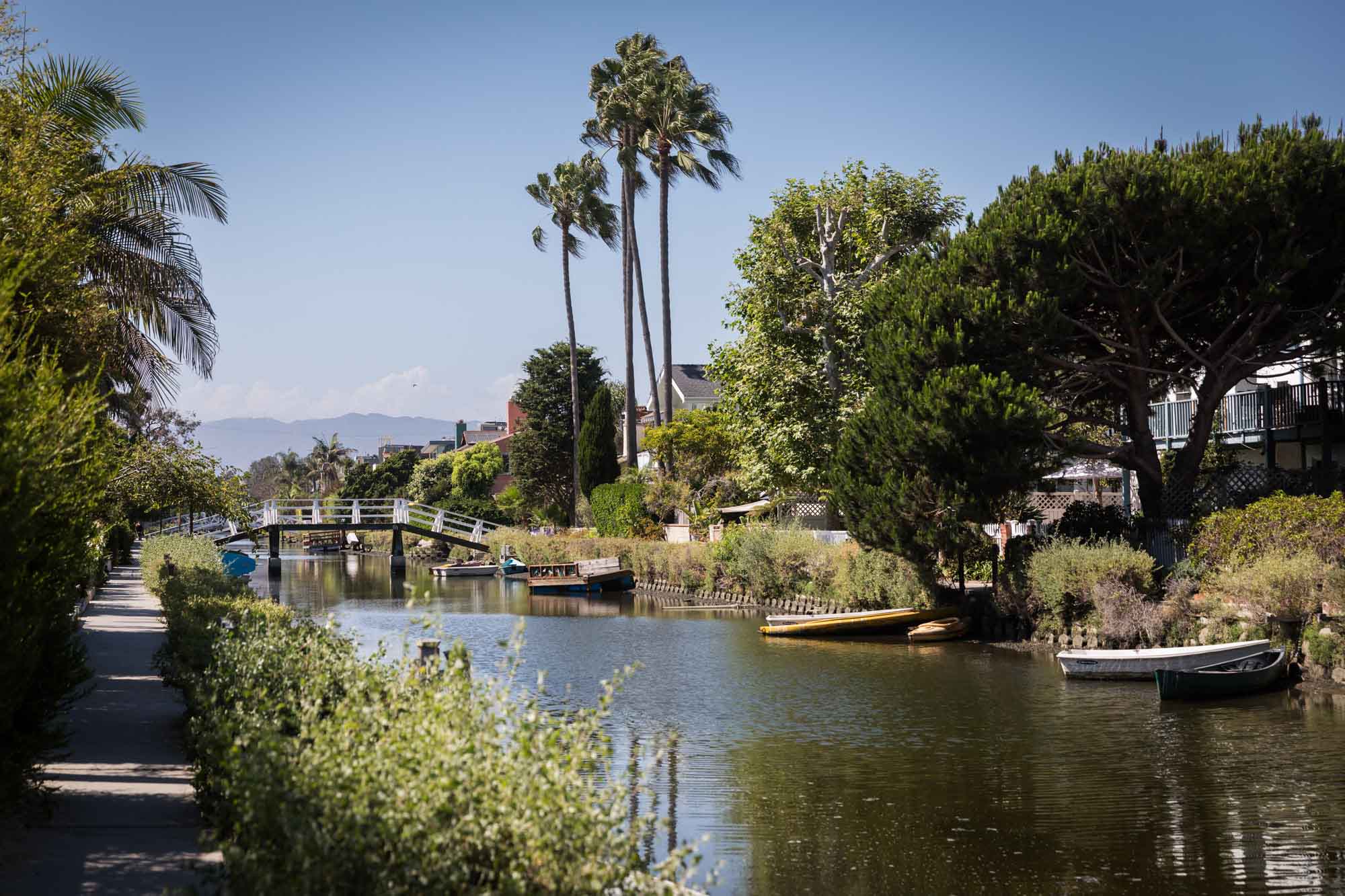 White footbridge over a canal with boats, trees, and small docks on the side along the Venice Canals in Los Angeles for an article on Venice Canals engagement photos