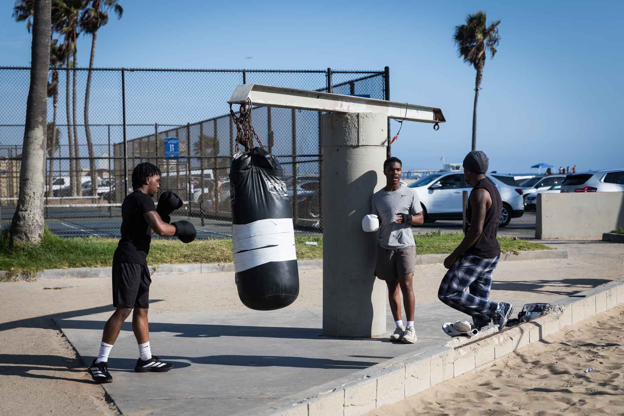 African American man wearing boxing gloves punching a boxing bag with two other African American men standing nearby at Venice Beach for an article on Venice Canals engagement photos