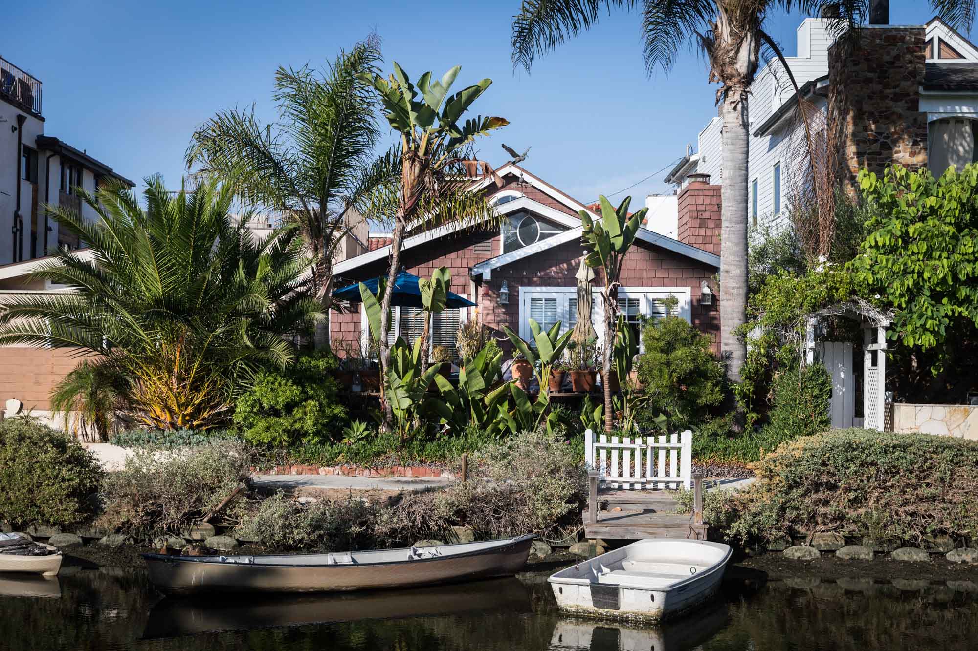 Wooden-shingle home with palm trees and wooden fence in front and two rowboats in the canal in the water for an article on Venice Canals engagement photos