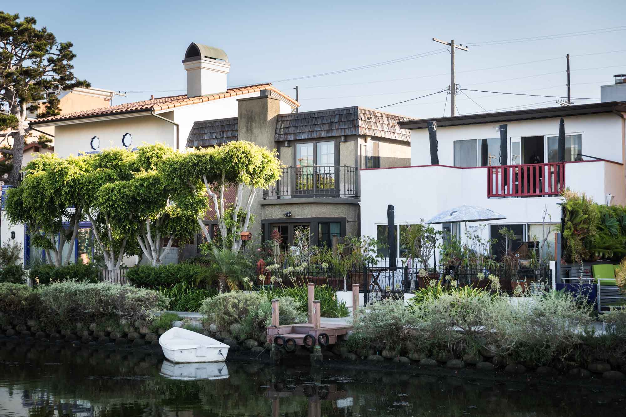Three different style homes along the side of the Venice Canals with a white rowboat in the water beside a small dock for an article on Venice Canals engagement photos