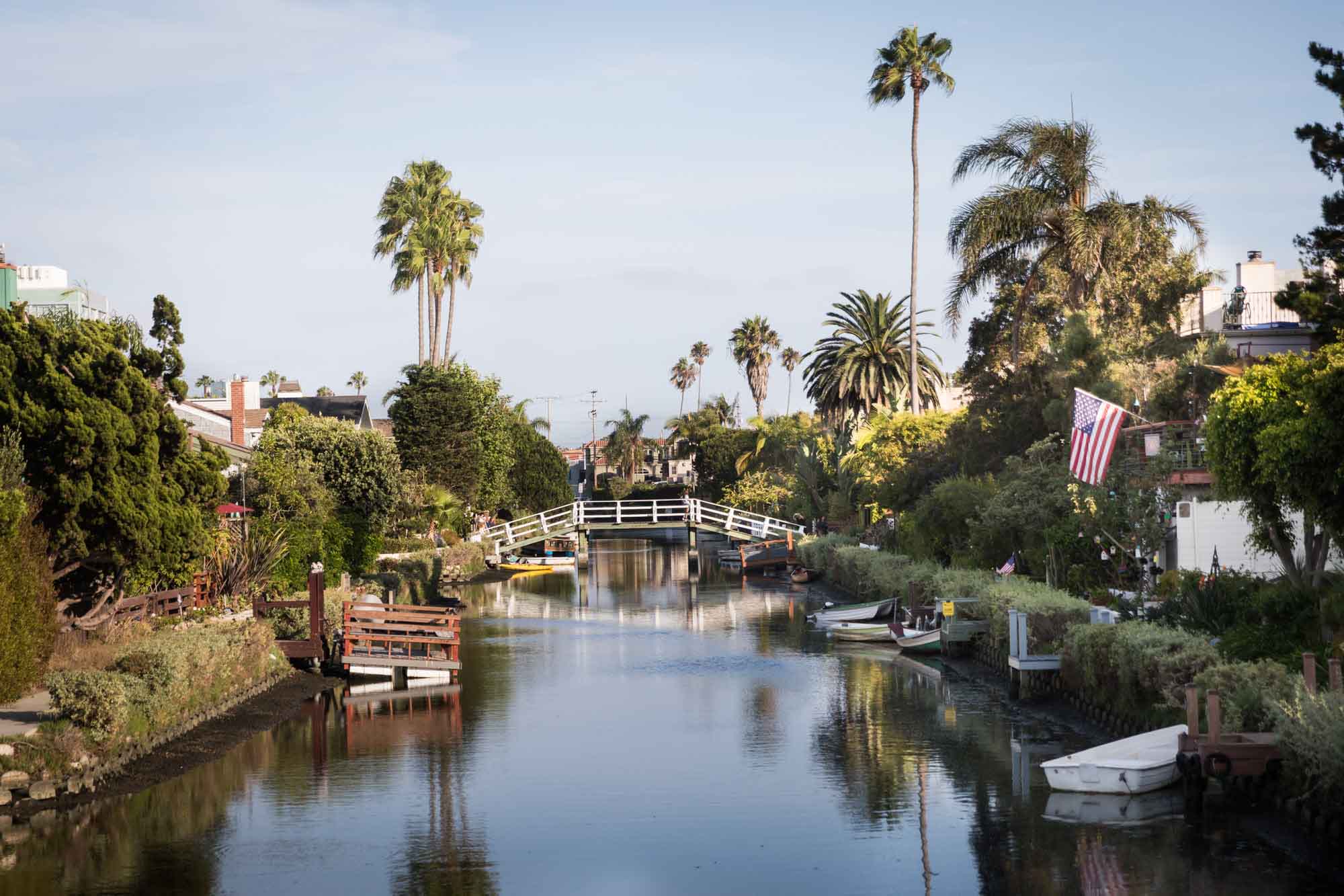 White footbridge over a canal with boats, trees, and small docks on the side along the Venice Canals in Los Angeles for an article on Venice Canals engagement photos