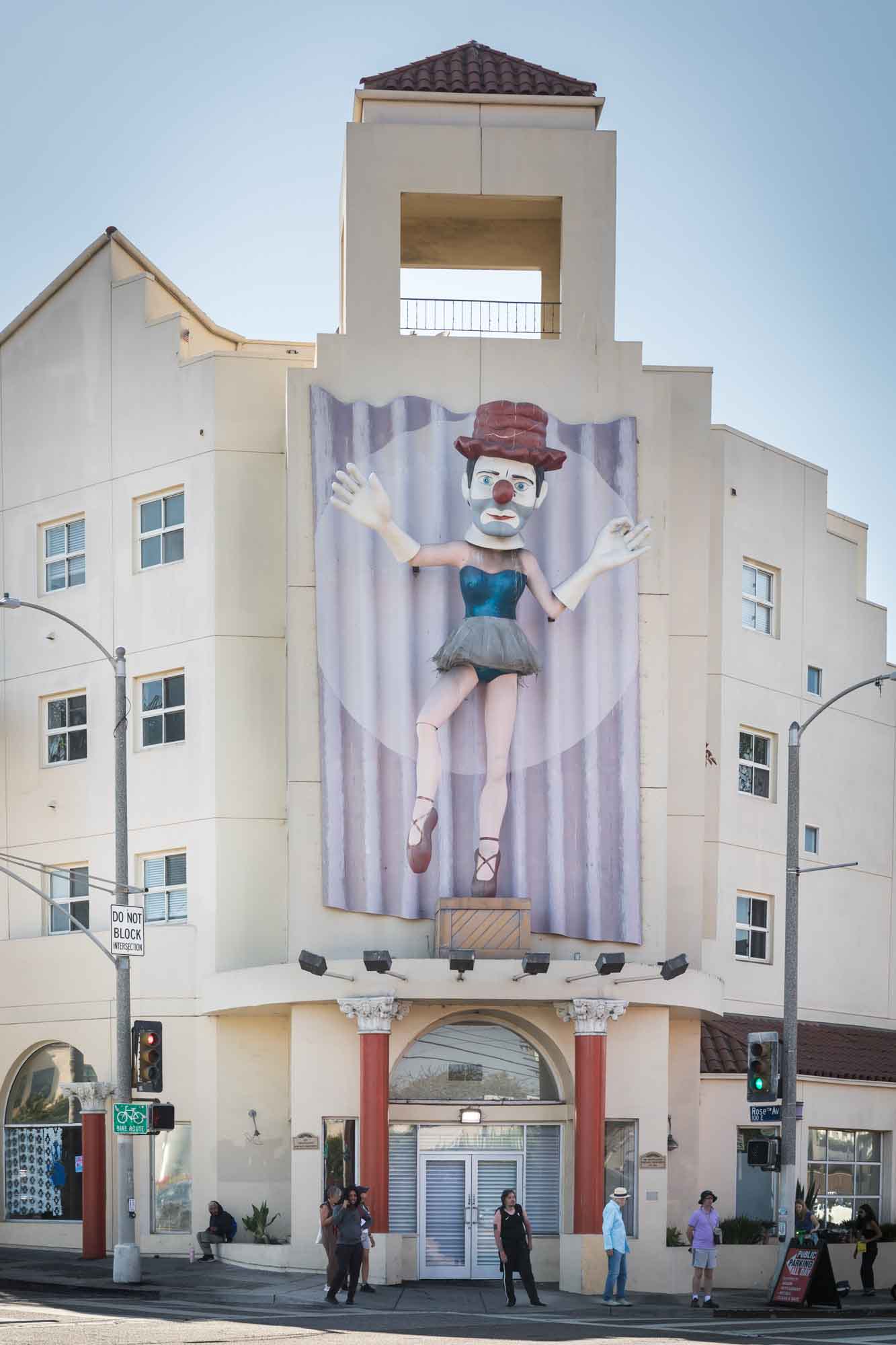 Ballerina Clown public art piece above a beige building with red columns below and people waiting to cross the street in Venice for an article on Venice Canals engagement photos