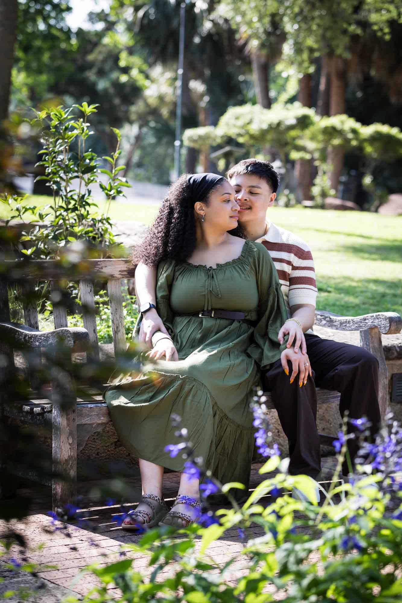 Man hugging woman while sitting together on wooden bench in a garden for an article on Venice Canals engagement photos