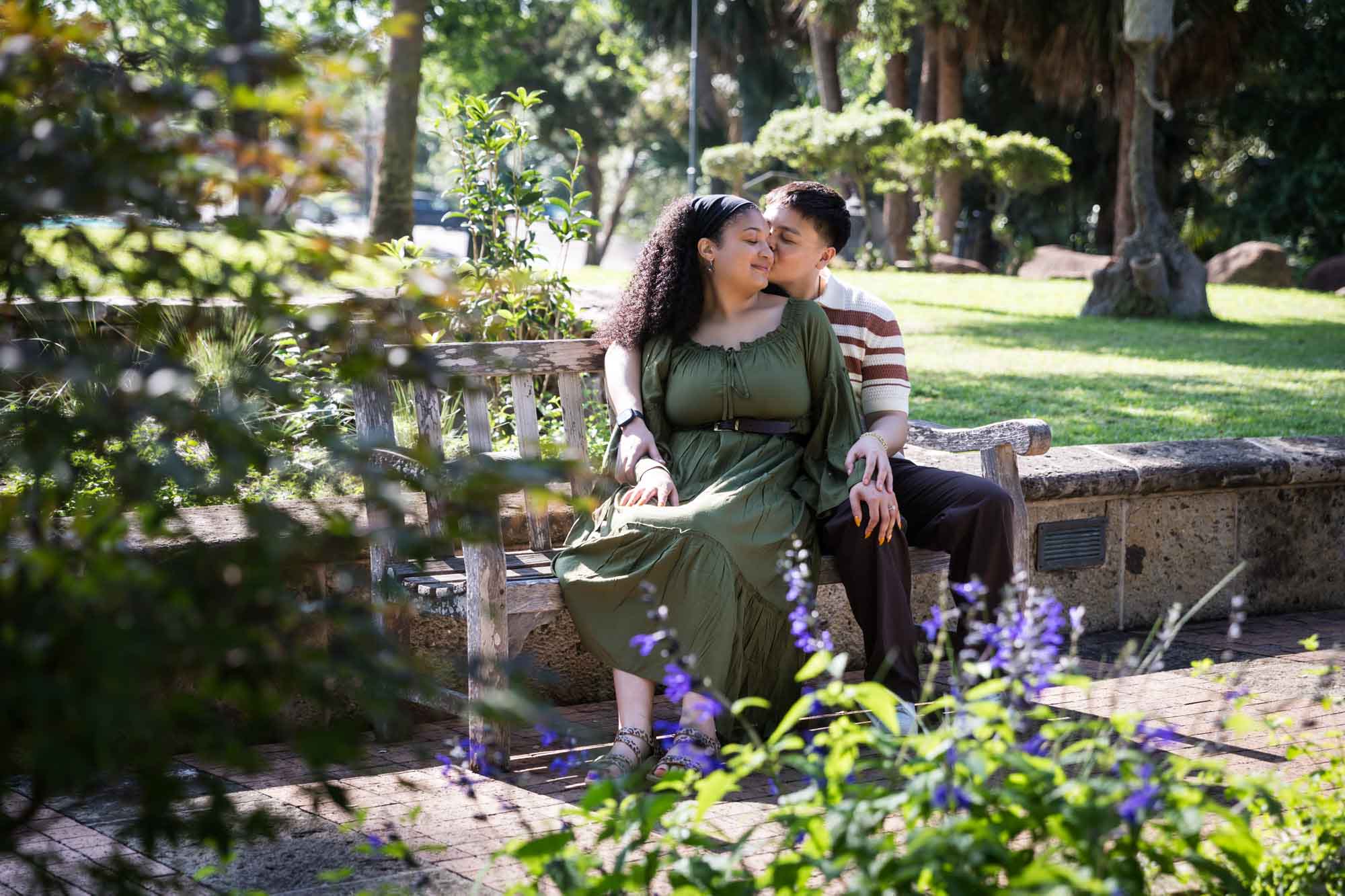 Man kissing woman while sitting together on wooden bench in a garden for an article on Venice Canals engagement photos