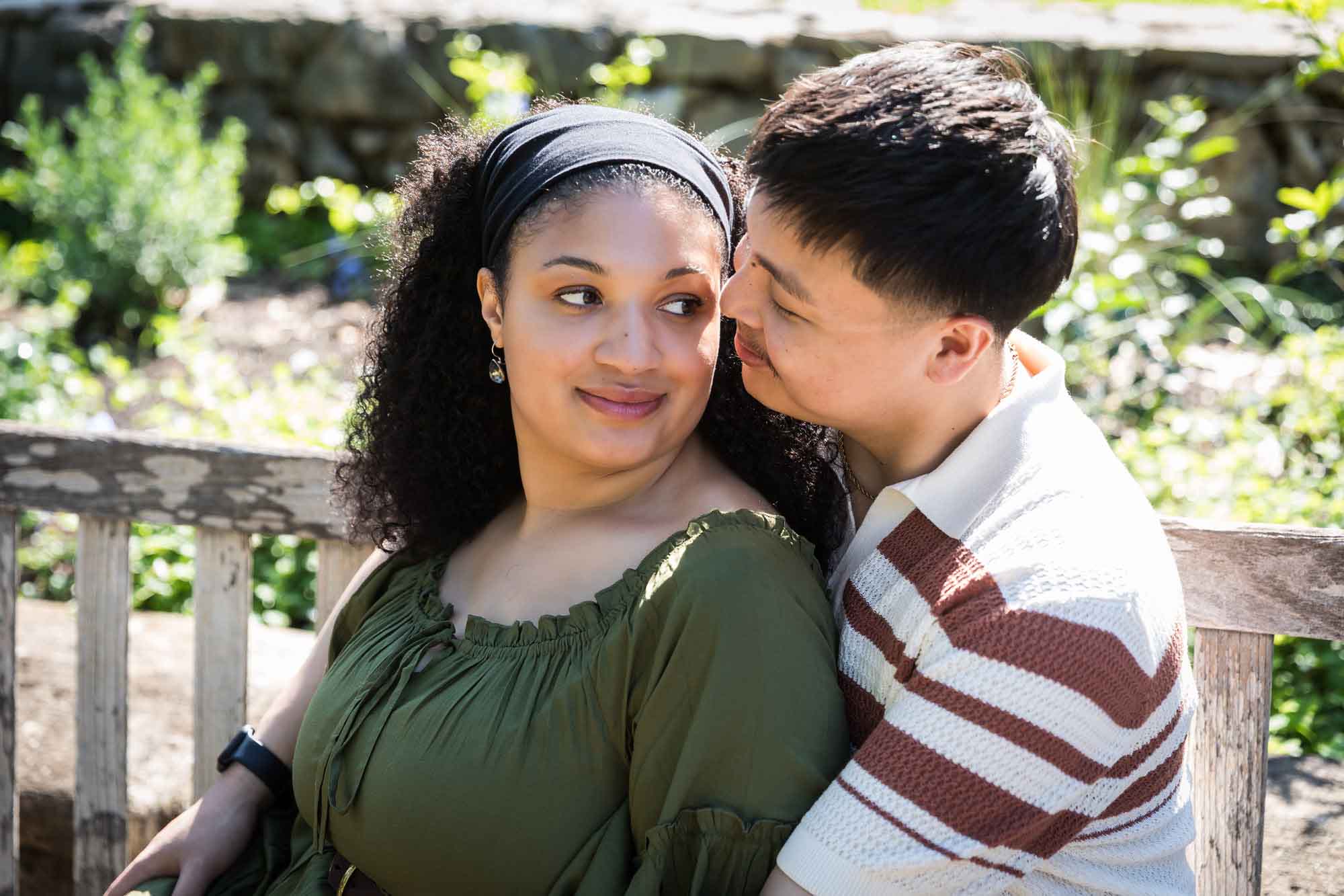 Man hugging woman while sitting together on wooden bench in a garden for an article on Venice Canals engagement photos