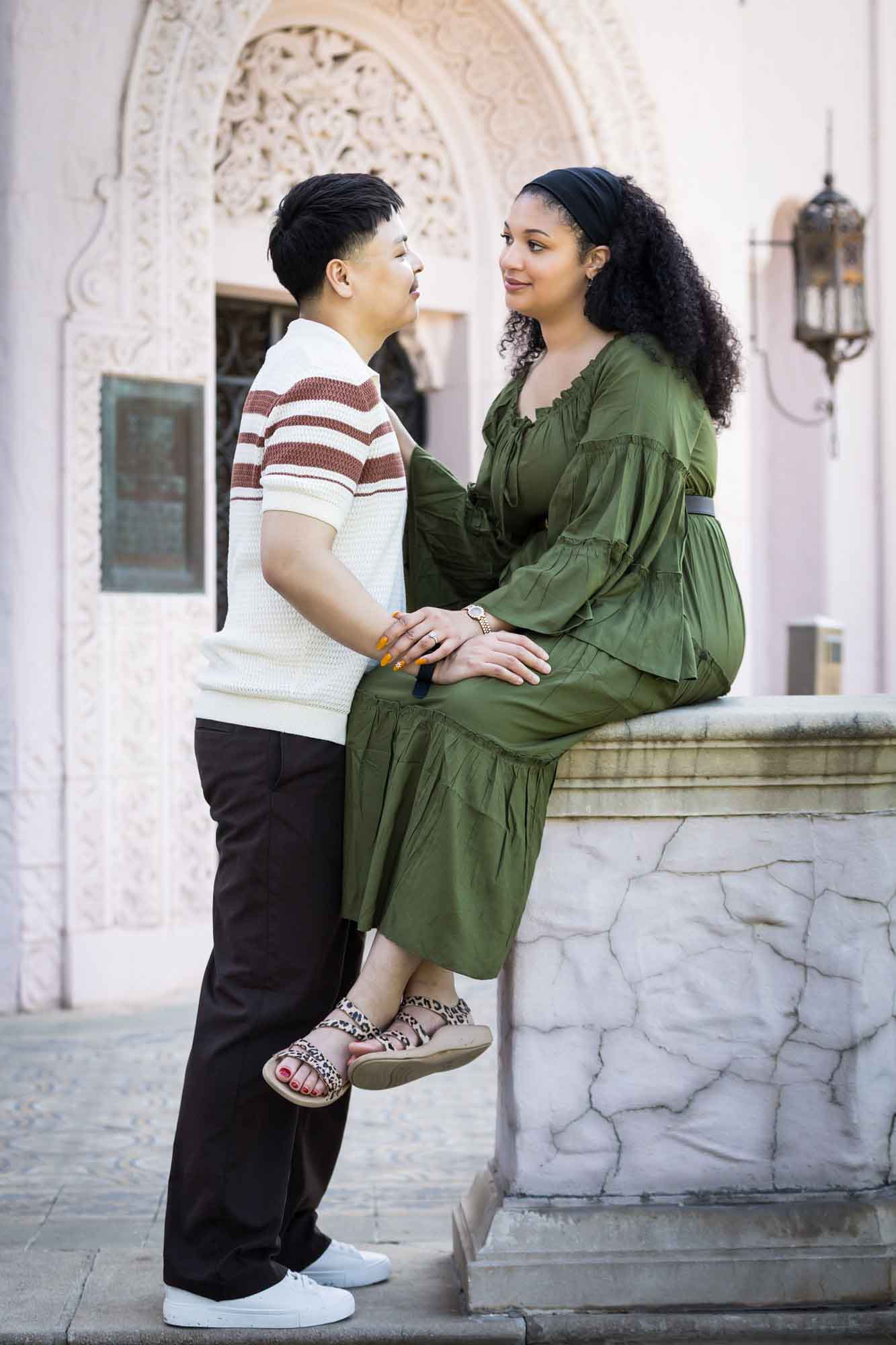 Woman in green dress sitting on stone pedestal holding arm of man in front of intricate stone home for an article on Venice Canals engagement photos