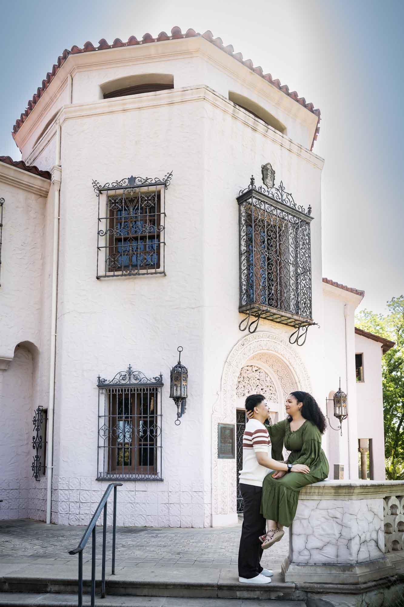 Woman in green dress sitting on stone pedestal holding arm of man in front of intricate stone home for an article on Venice Canals engagement photos