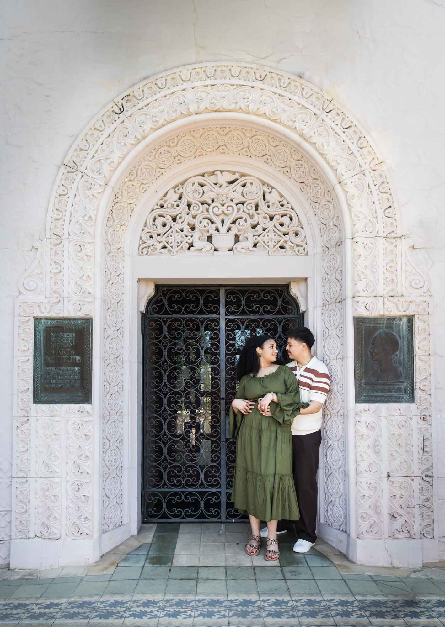 Man hugging woman wearing green dress in front of intricately carved stone arch doorway with wrought iron door for an article on Venice Canals engagement photos